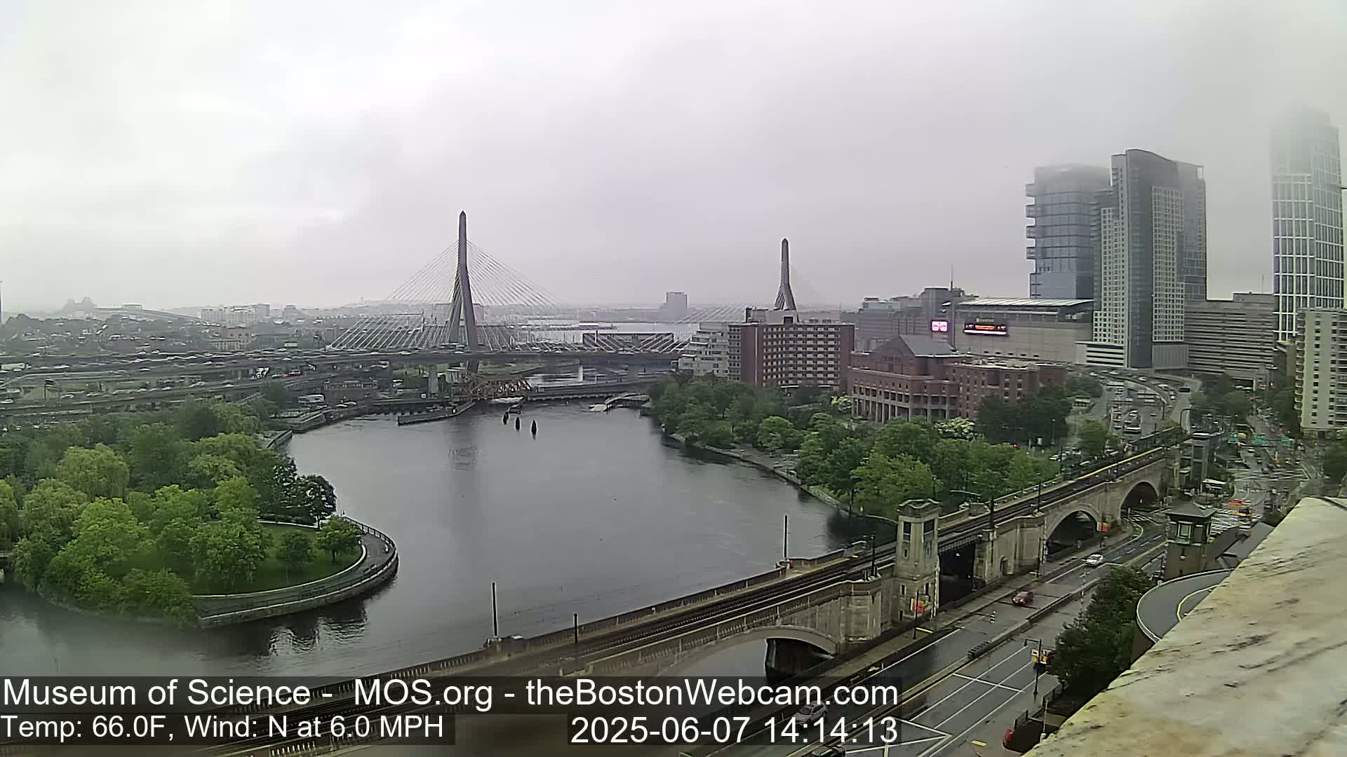 A foggy cityscape shows a river with two bridges, one a cable-stayed bridge, the other an arched bridge, with buildings and lush green spaces along the riverbanks.