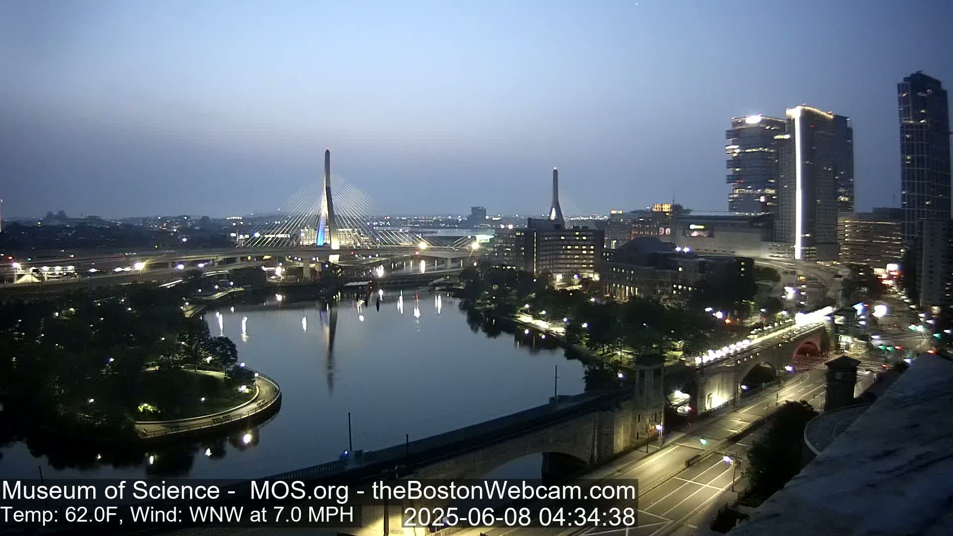 A nighttime cityscape with two cable-stayed bridges over a dark river, lit city streets, and tall buildings under a twilight sky.
