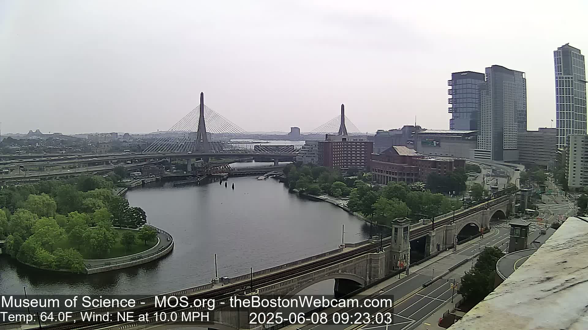 A city waterfront scene on a cloudy day shows two cable-stayed bridges spanning a river, with green parkland, buildings, and roadways visible.