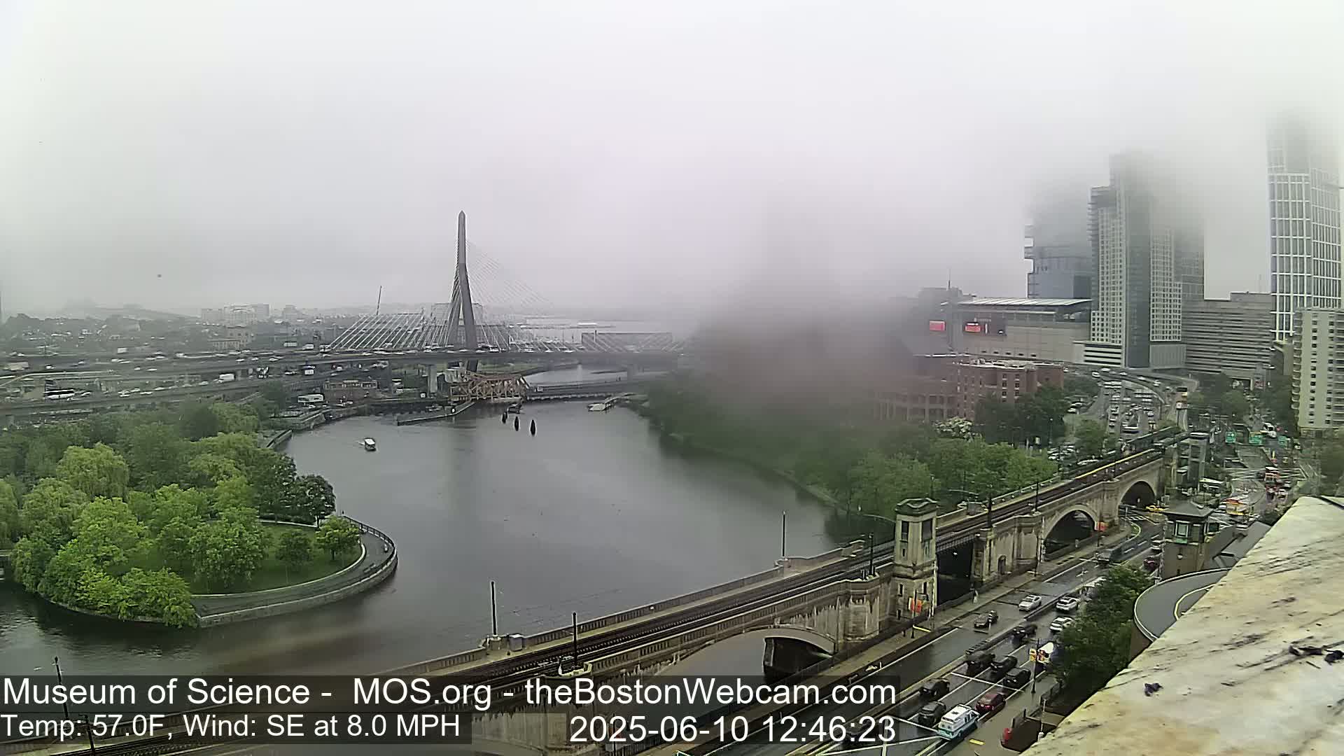 A foggy cityscape shows a river with a bridge and a cable-stayed bridge in the distance, with city buildings and roadways visible through the mist.