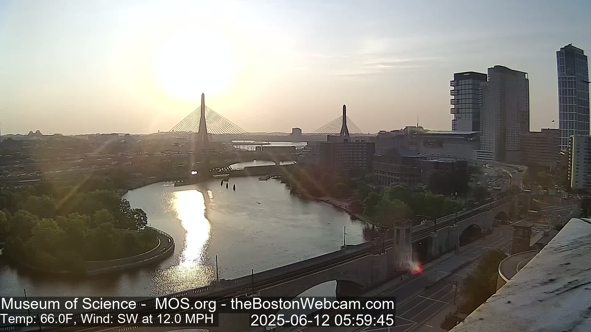 A sunrise illuminates two cable-stayed bridges spanning a river, with city buildings and a roadway visible, under a partly sunny sky with a southwest wind at 12 mph.