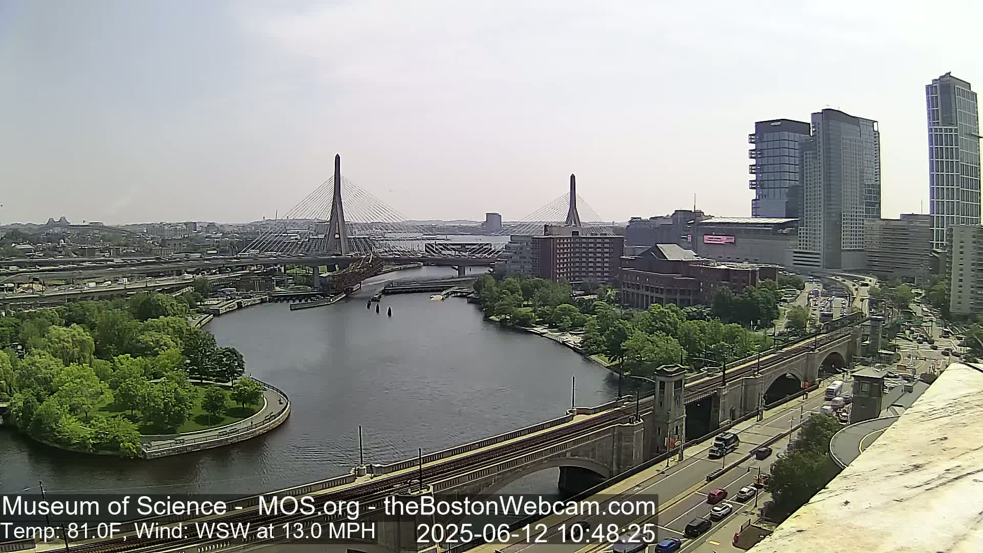 A sunny city view shows two cable-stayed bridges spanning a river, with modern high-rise buildings, green spaces, and roadways in the background.
