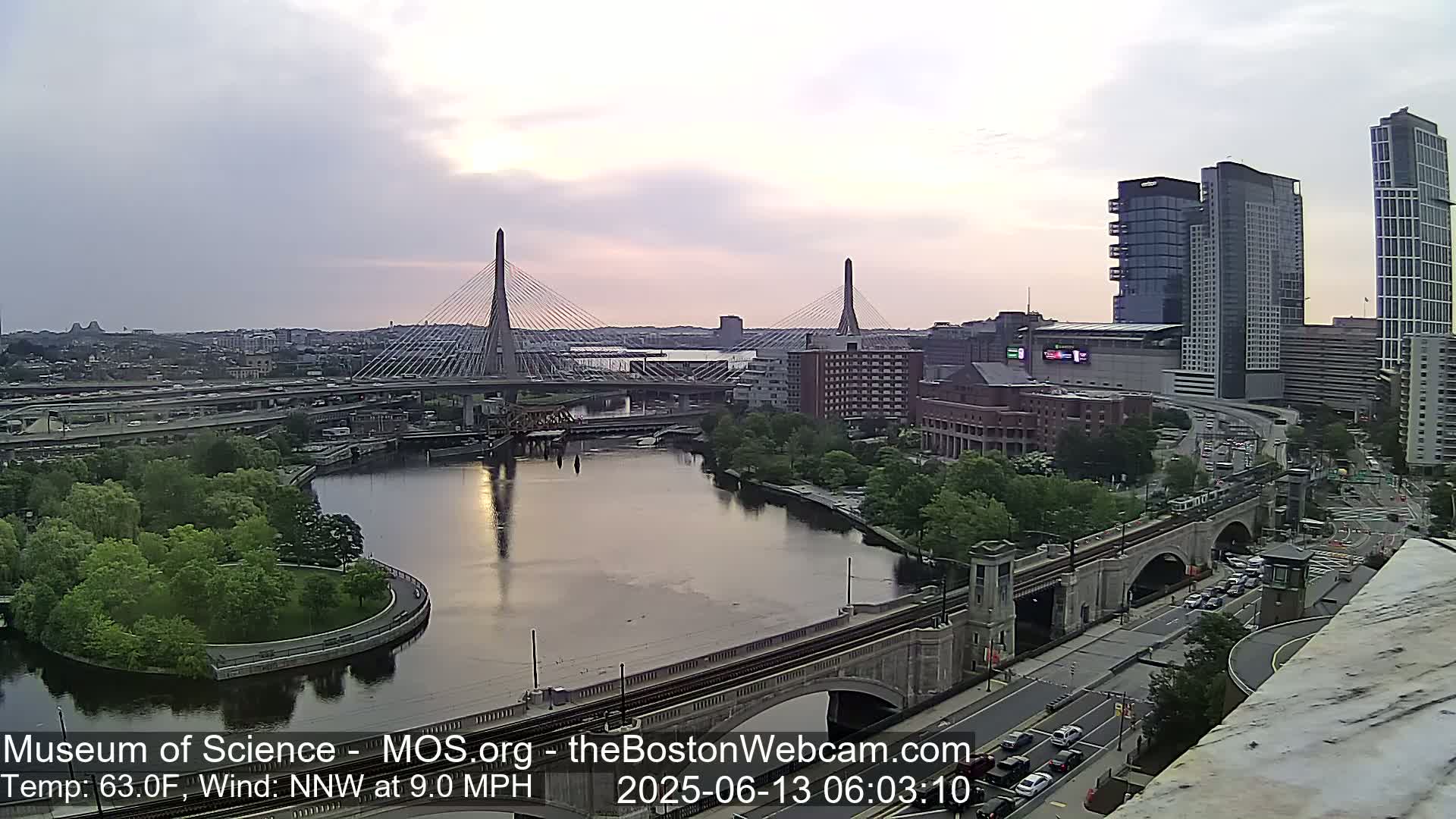 A city skyline with two cable-stayed bridges over a river, under a partly cloudy sky at sunrise.