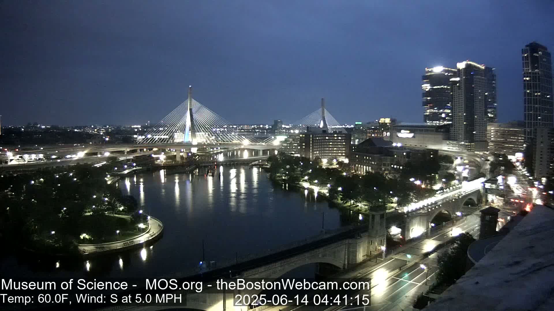 A nighttime cityscape shows two cable-stayed bridges spanning a river, with city lights reflecting on the water under a dark blue sky; the wind is from the south at 5 mph.