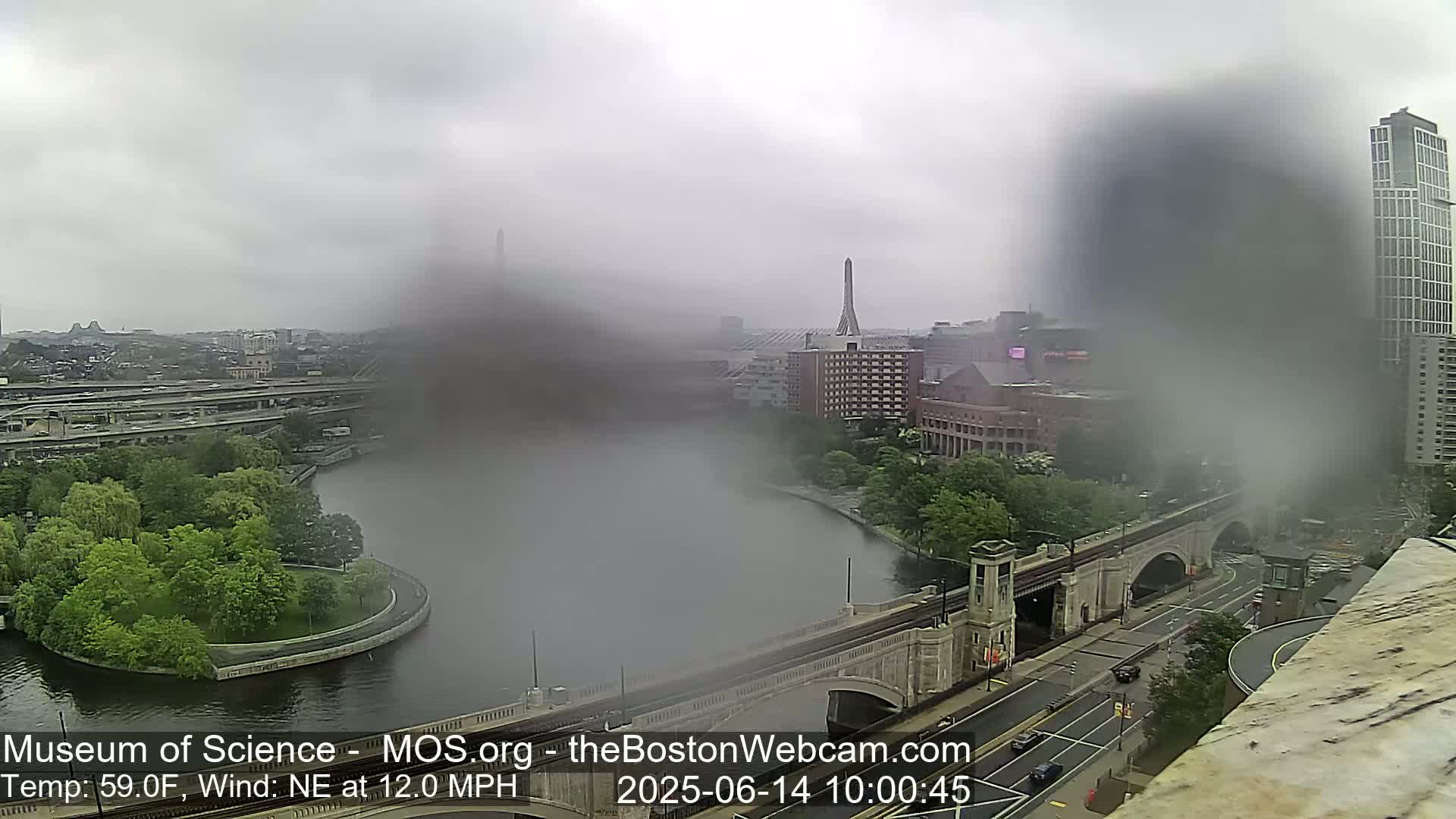 A hazy, rainy view of a city with a river, bridge, and parkland, viewed from a high vantage point.