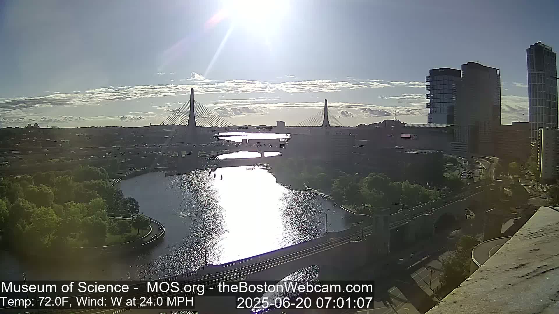 A sunny, windy day reveals two cable-stayed bridges spanning a waterway, with city buildings and green spaces visible in the background.