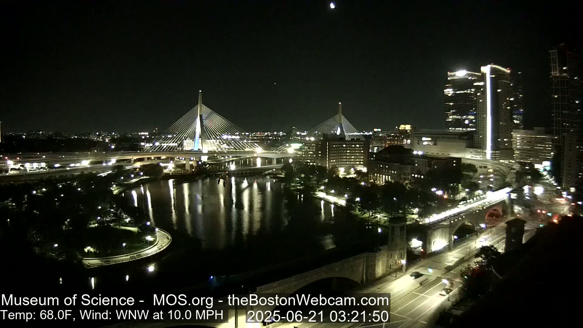 A nighttime view of two illuminated cable-stayed bridges spanning a dark river, with city lights and buildings visible in the background under a clear sky.