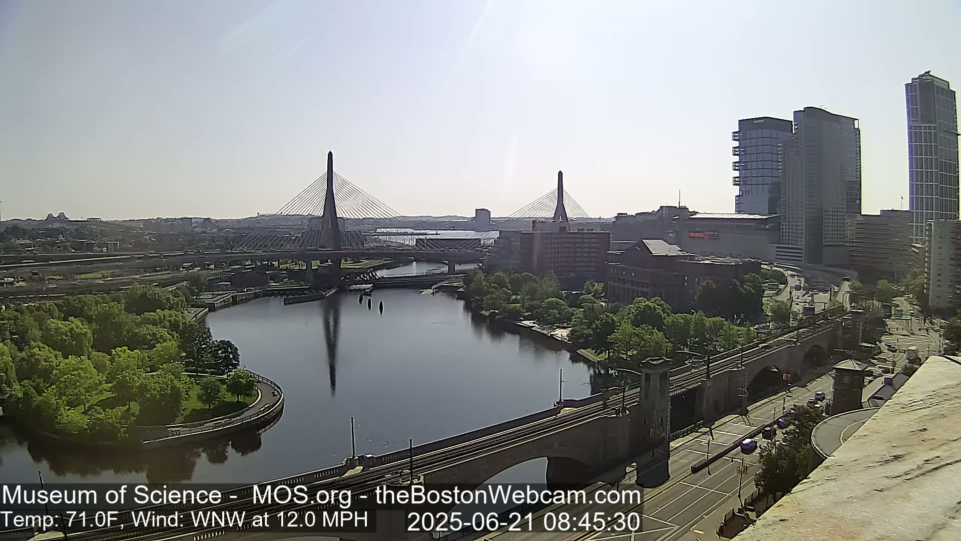 A sunny cityscape view shows two cable-stayed bridges spanning a river, with green parkland, buildings, and a train trestle visible.