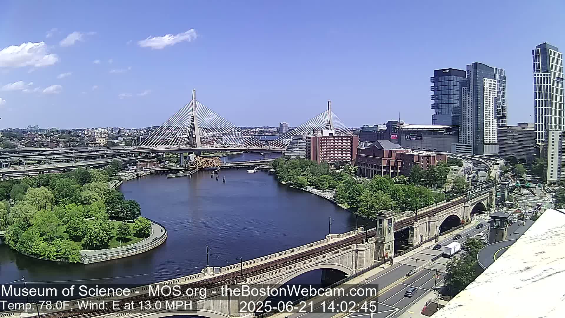A sunny cityscape features two cable-stayed bridges spanning a river, with a roadway and several buildings visible.