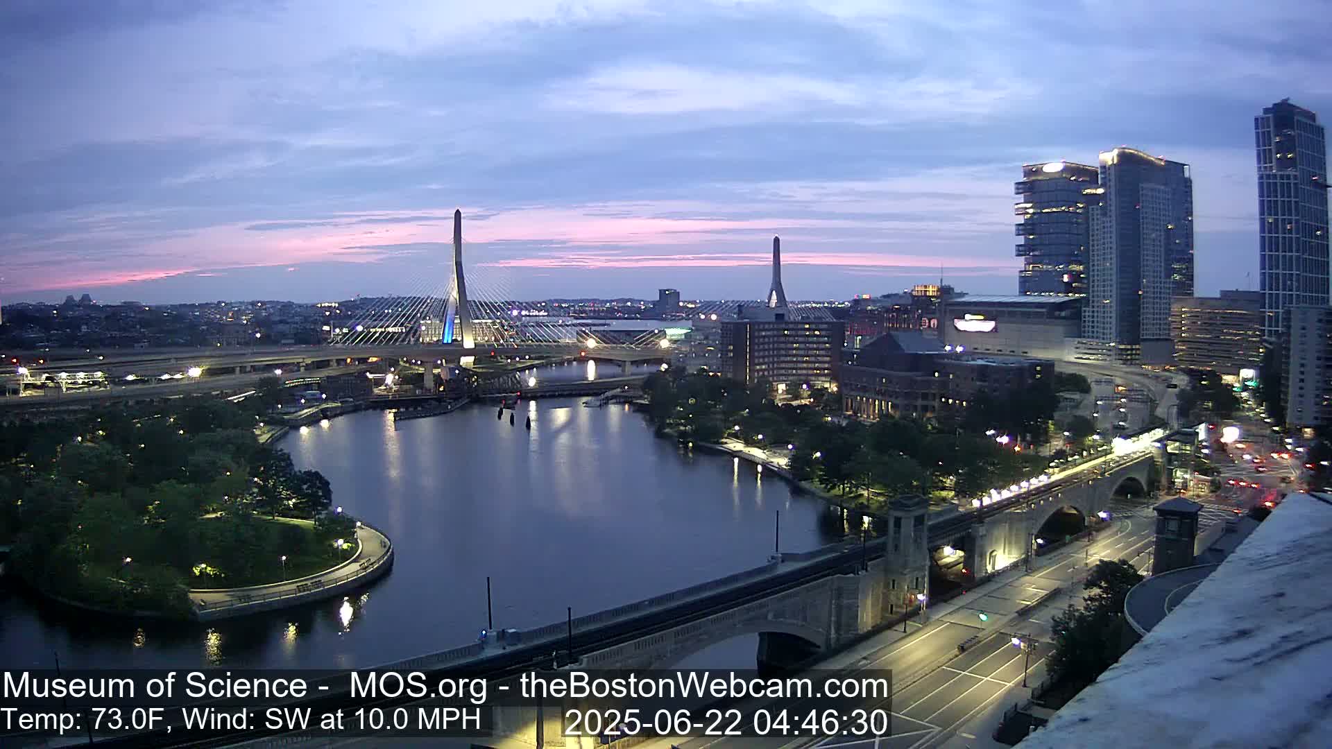 A twilight cityscape shows two cable-stayed bridges over a dark river, with city buildings and a park along the waterfront under a mostly clear sky.