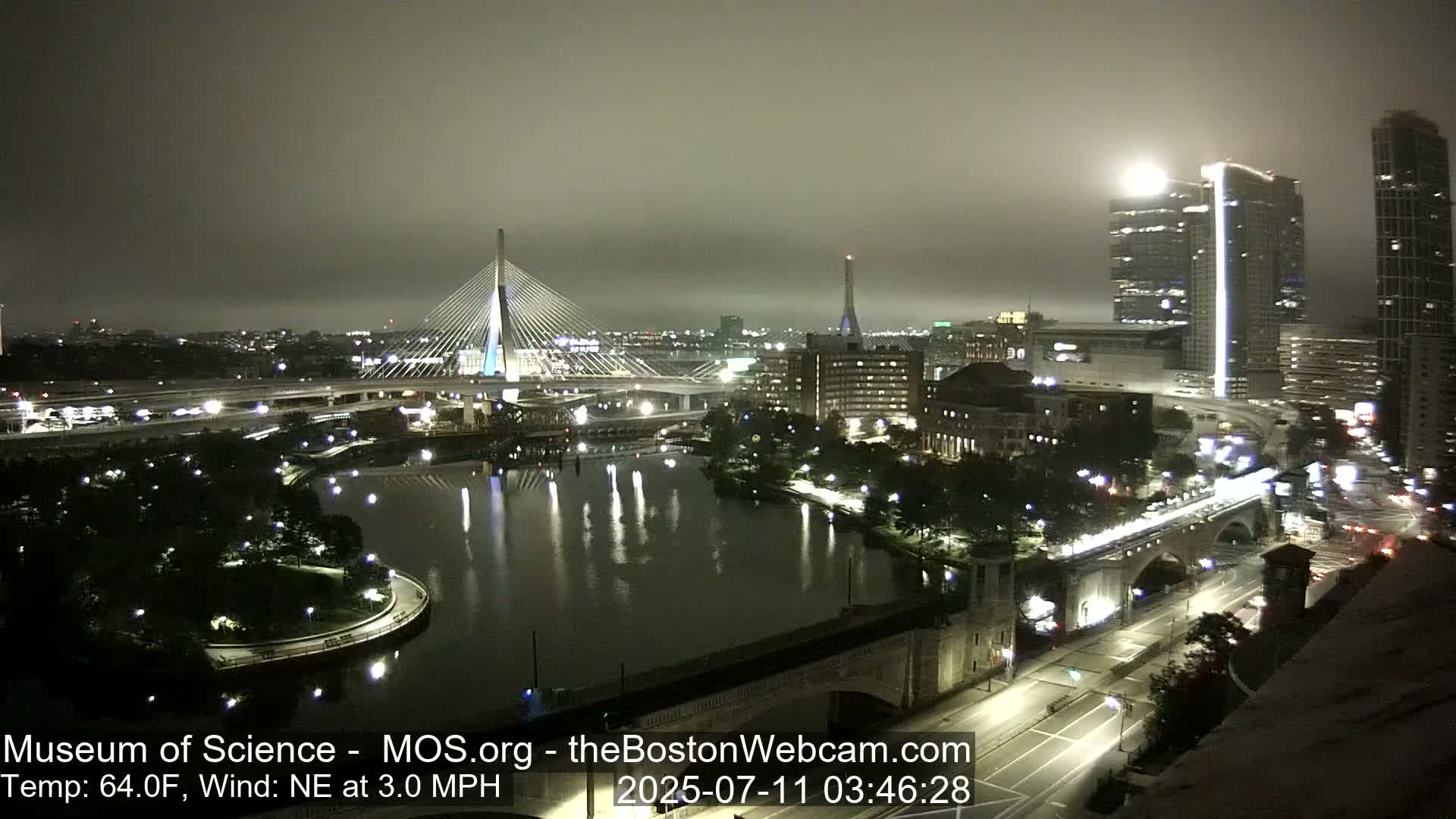 A nighttime cityscape with a bridge and river, shrouded in fog under a dark sky.