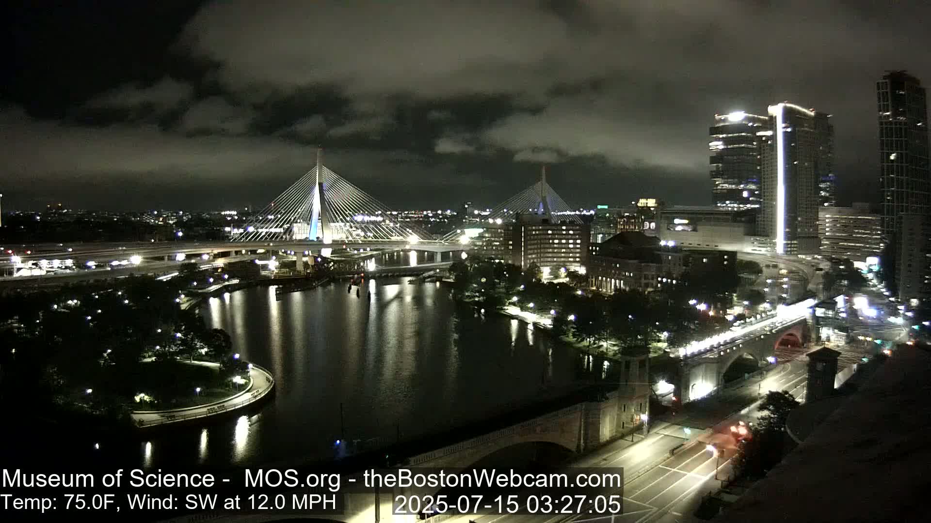 A nighttime cityscape with two illuminated cable-stayed bridges spanning a dark river, under a partly cloudy sky, with a southwest wind at 12 mph and a temperature of 75 degrees Fahrenheit.
