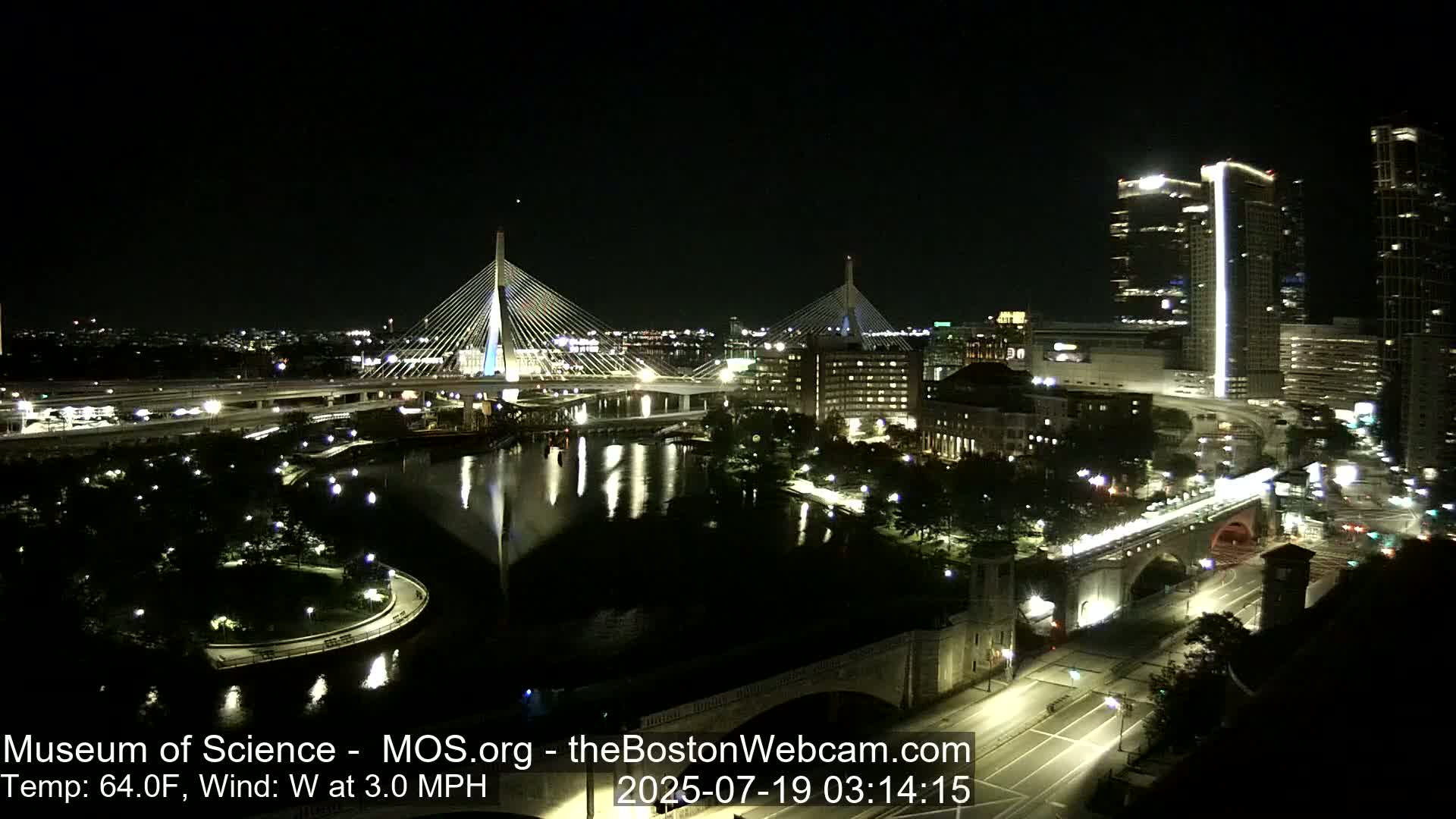 A nighttime view of two cable-stayed bridges spanning a dark river, surrounded by city lights and a calm body of water.