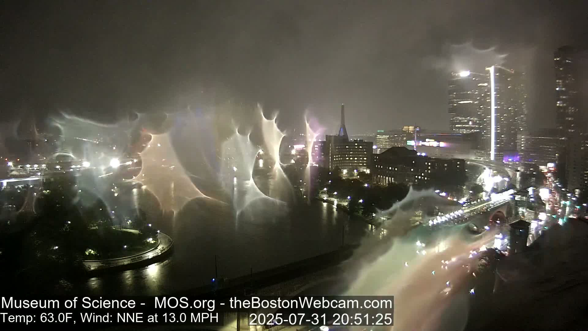 A nighttime view of a city skyline with a river, illuminated by city lights and distorted by heavy rain and wind.