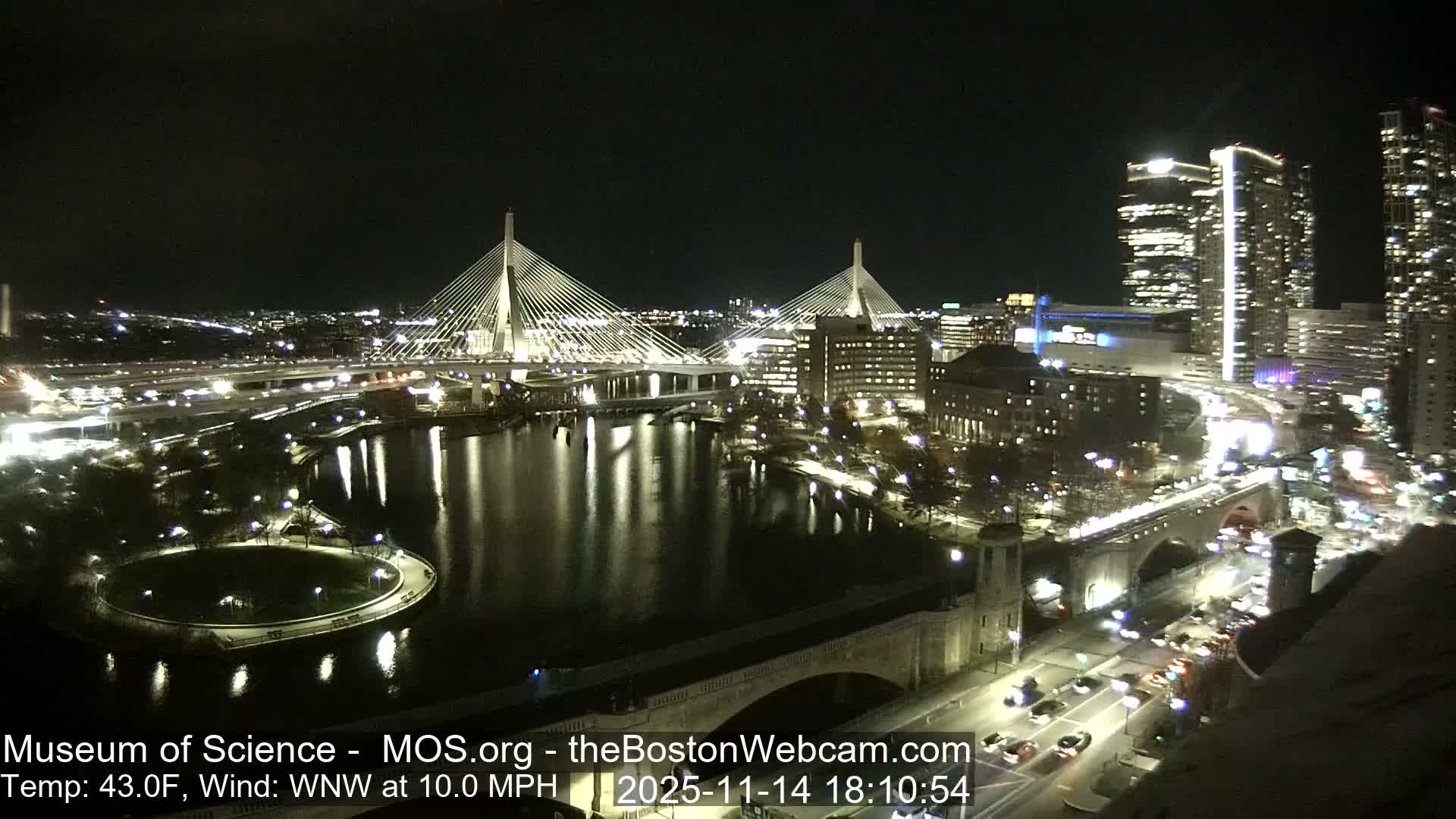 An illuminated cityscape is captured at night, showcasing two prominent cable-stayed bridges spanning a body of water, surrounded by bright buildings and active roads, under clear skies with a temperature of 43.0F and a WNW wind at 10.0 MPH.