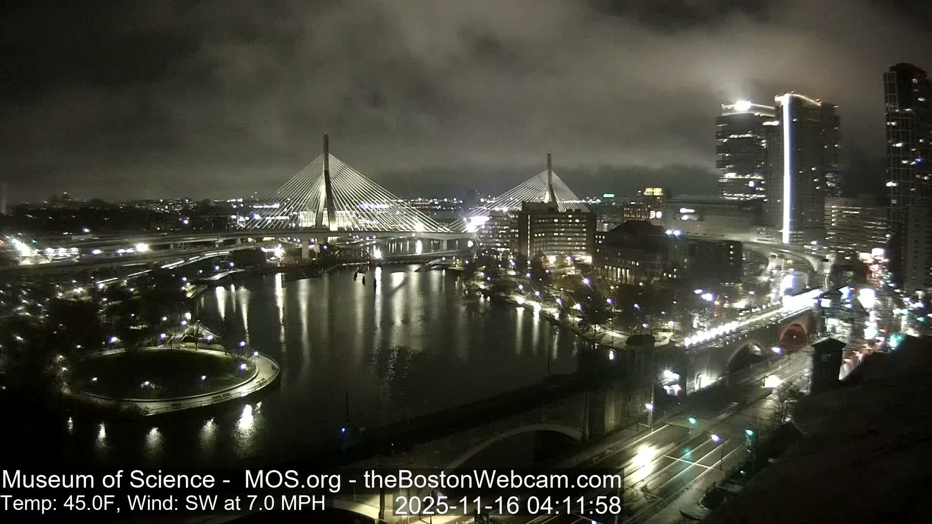 An illuminated cityscape at night features two prominent cable-stayed bridges spanning a body of water, surrounded by numerous lit buildings and roads, all viewed under an overcast sky.