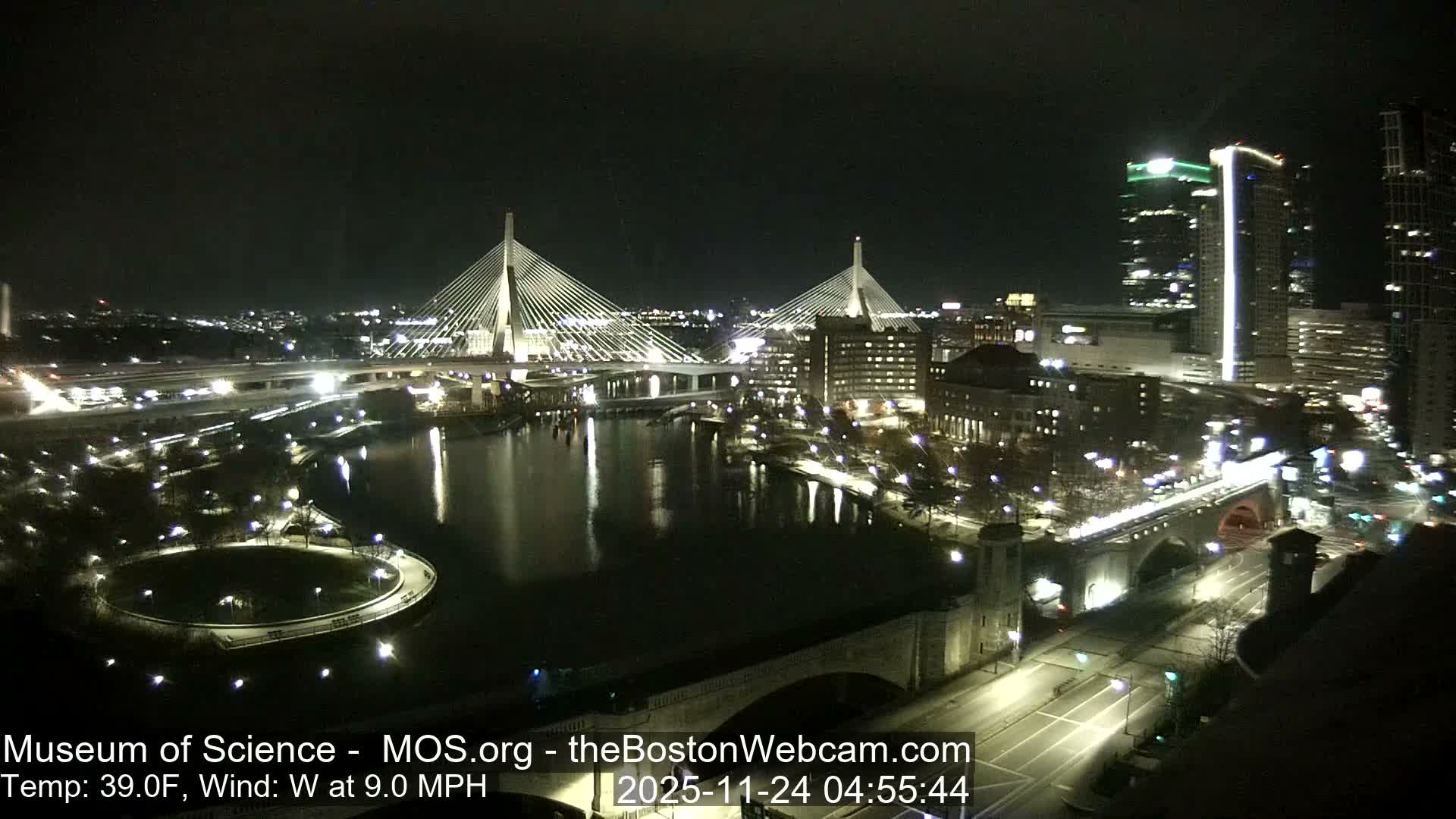 The image displays a sprawling cityscape at night, showcasing two brilliantly lit cable-stayed bridges arching over a dark river that reflects the surrounding urban lights and towering buildings, with a current temperature of 39.0F and a west wind blowing at 9.0 MPH.