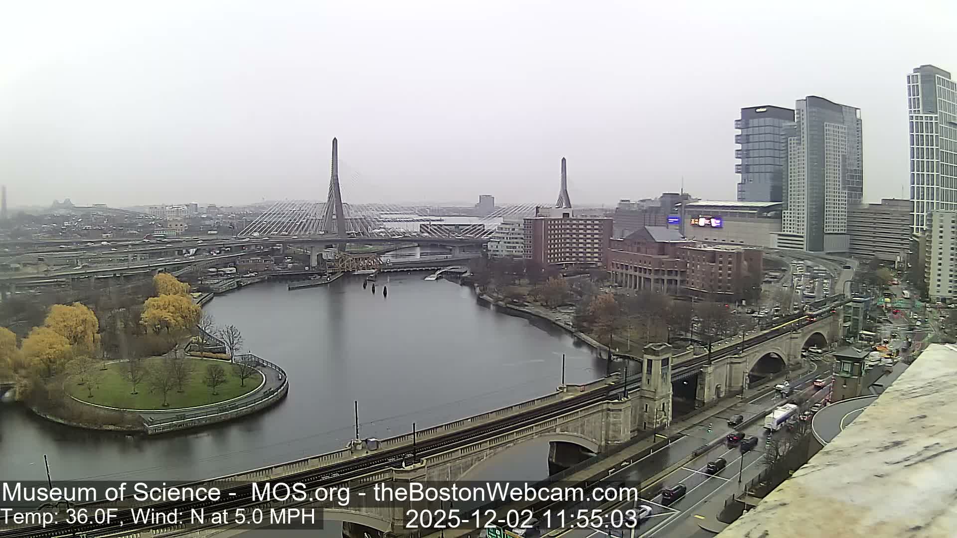 An elevated view reveals a sprawling city under an overcast sky, featuring a wide river spanned by multiple bridges, including two distinct cable-stayed bridges, extensive highway interchanges, numerous buildings, and scattered trees with fall foliage along the riverbanks.