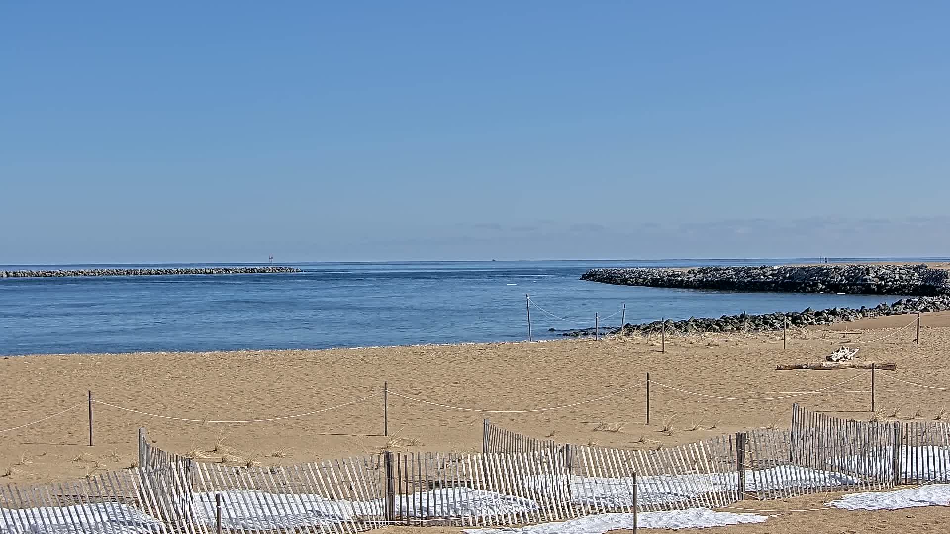 A sandy beach with a wooden fence and white coverings stretches towards a calm blue ocean under a clear sky.