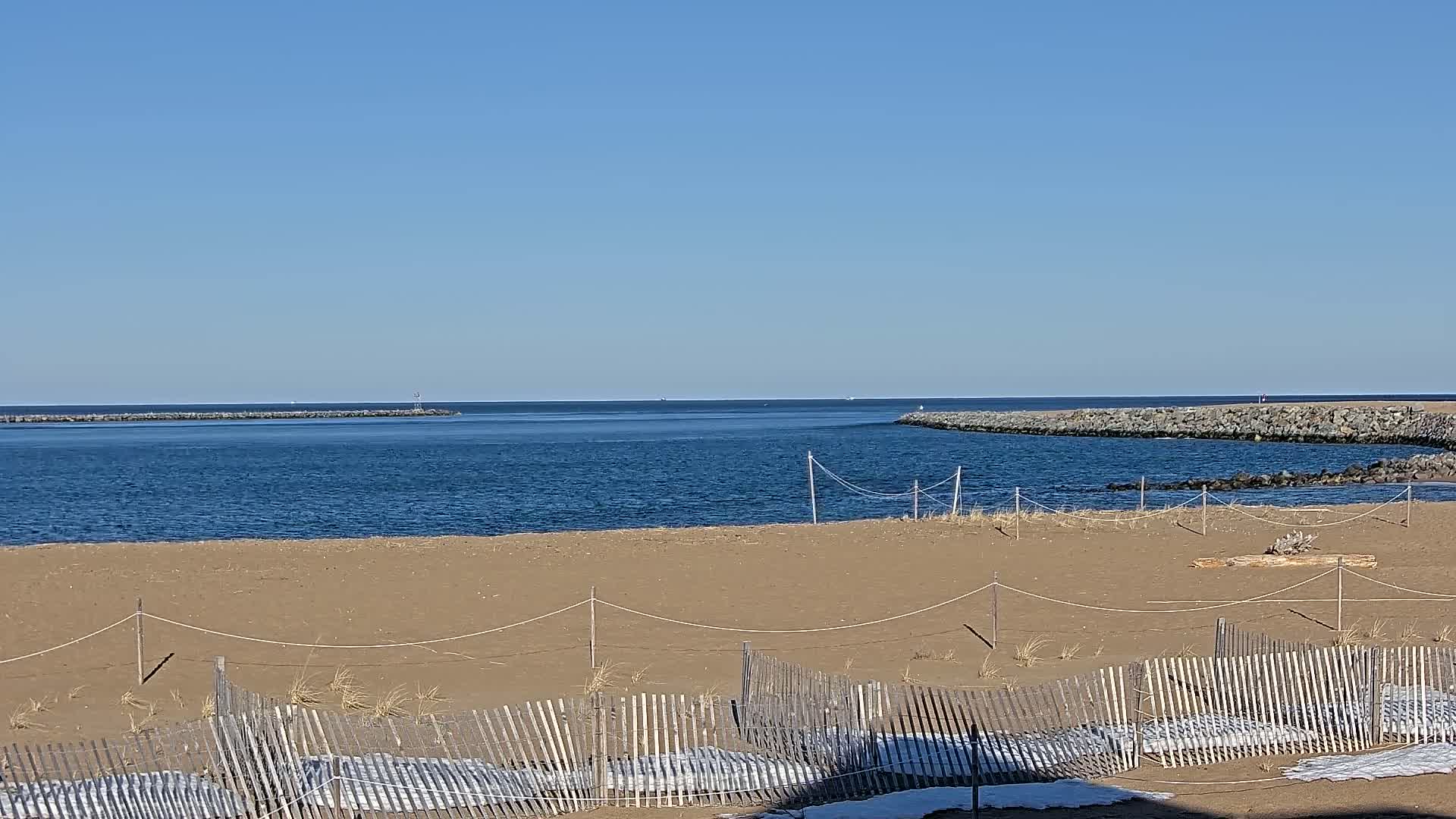 A sandy beach with a wooden fence partially covered in snow overlooks a calm blue ocean on a clear sunny day.
