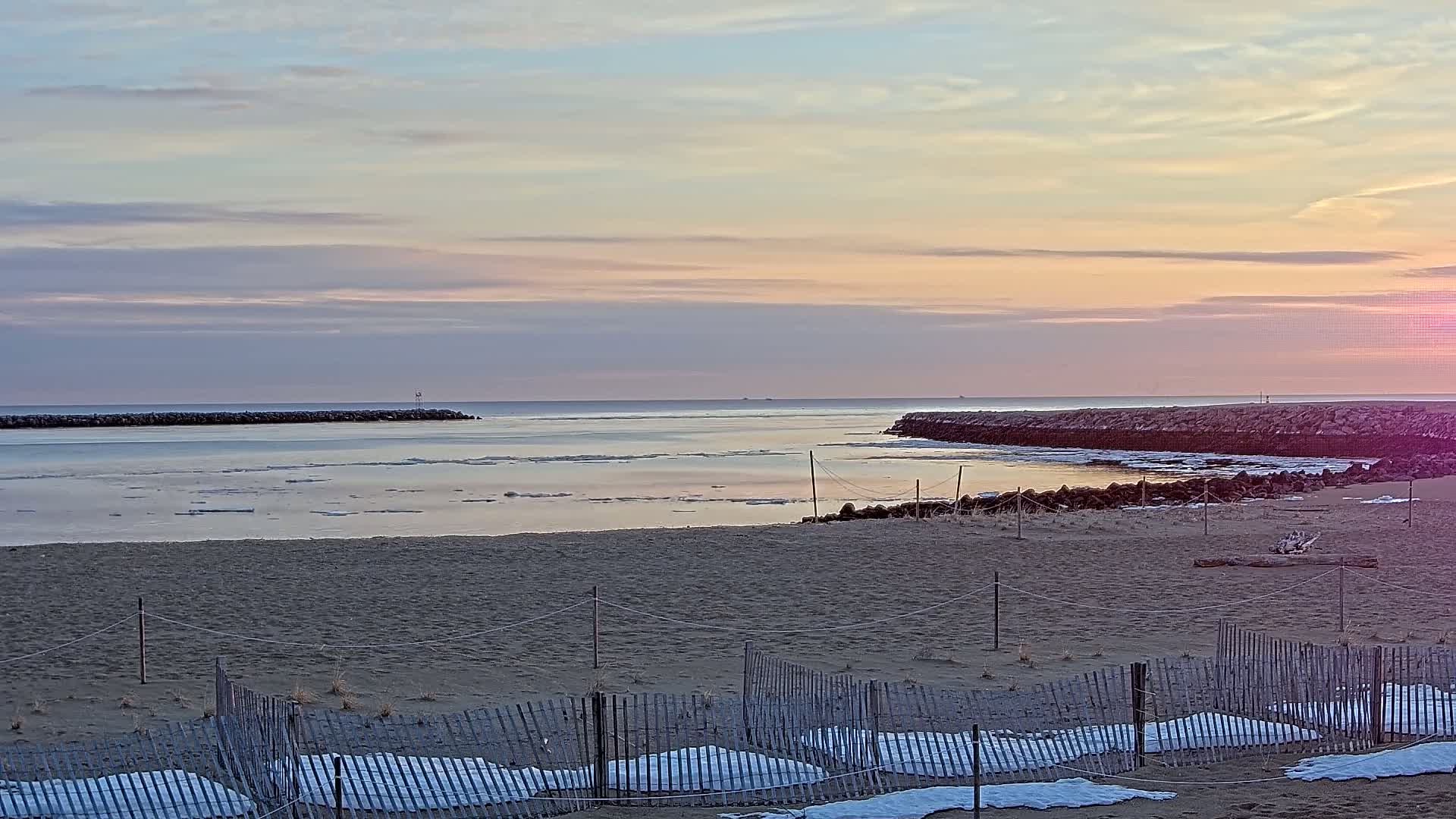 A sandy beach with partially snow-covered fences overlooks calm water with scattered ice floes under a pastel sunrise sky.
