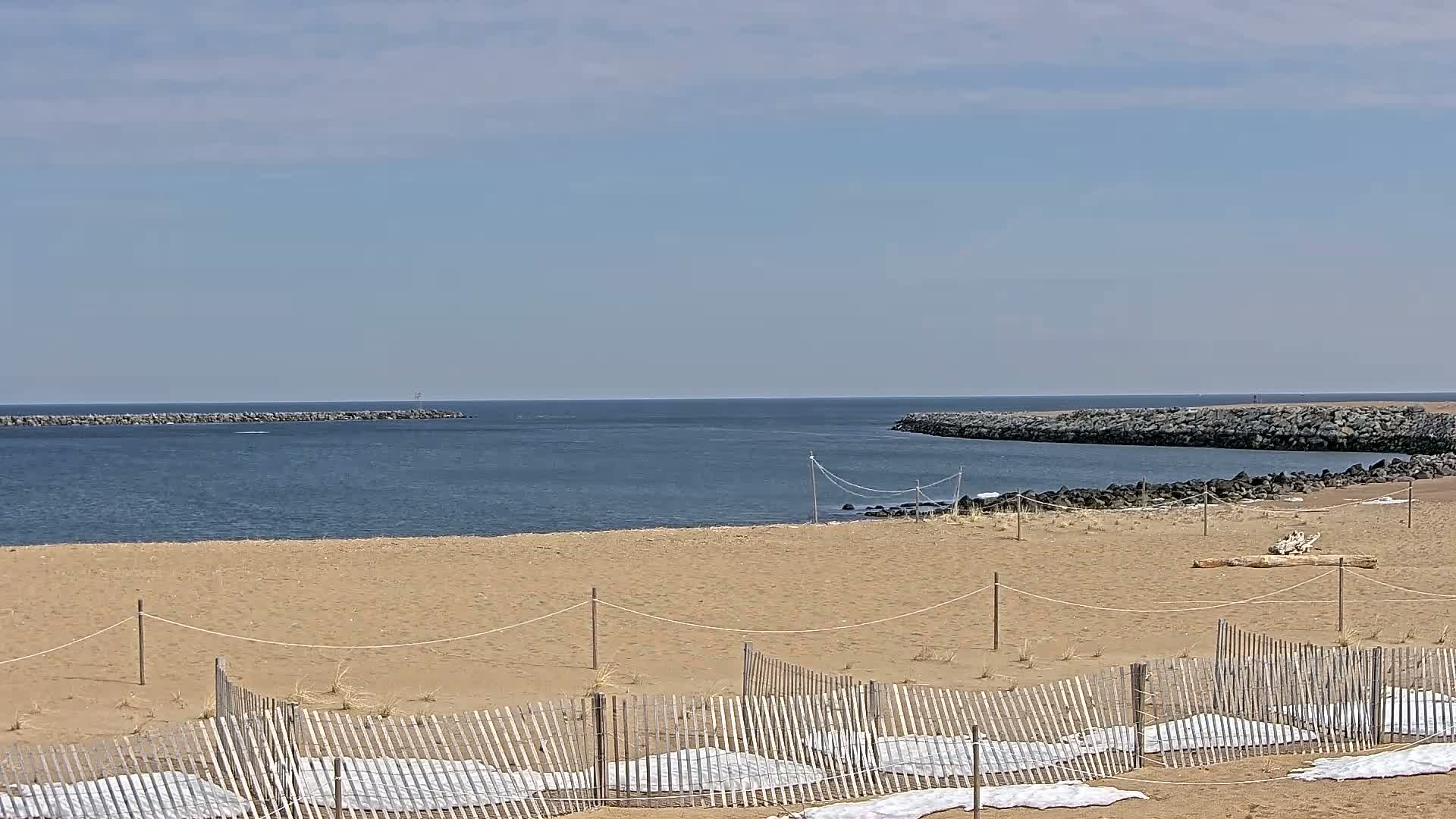 A sandy beach with a wooden fence and white coverings on the sand stretches toward a calm ocean inlet bordered by rock jetties under a mostly clear sky.