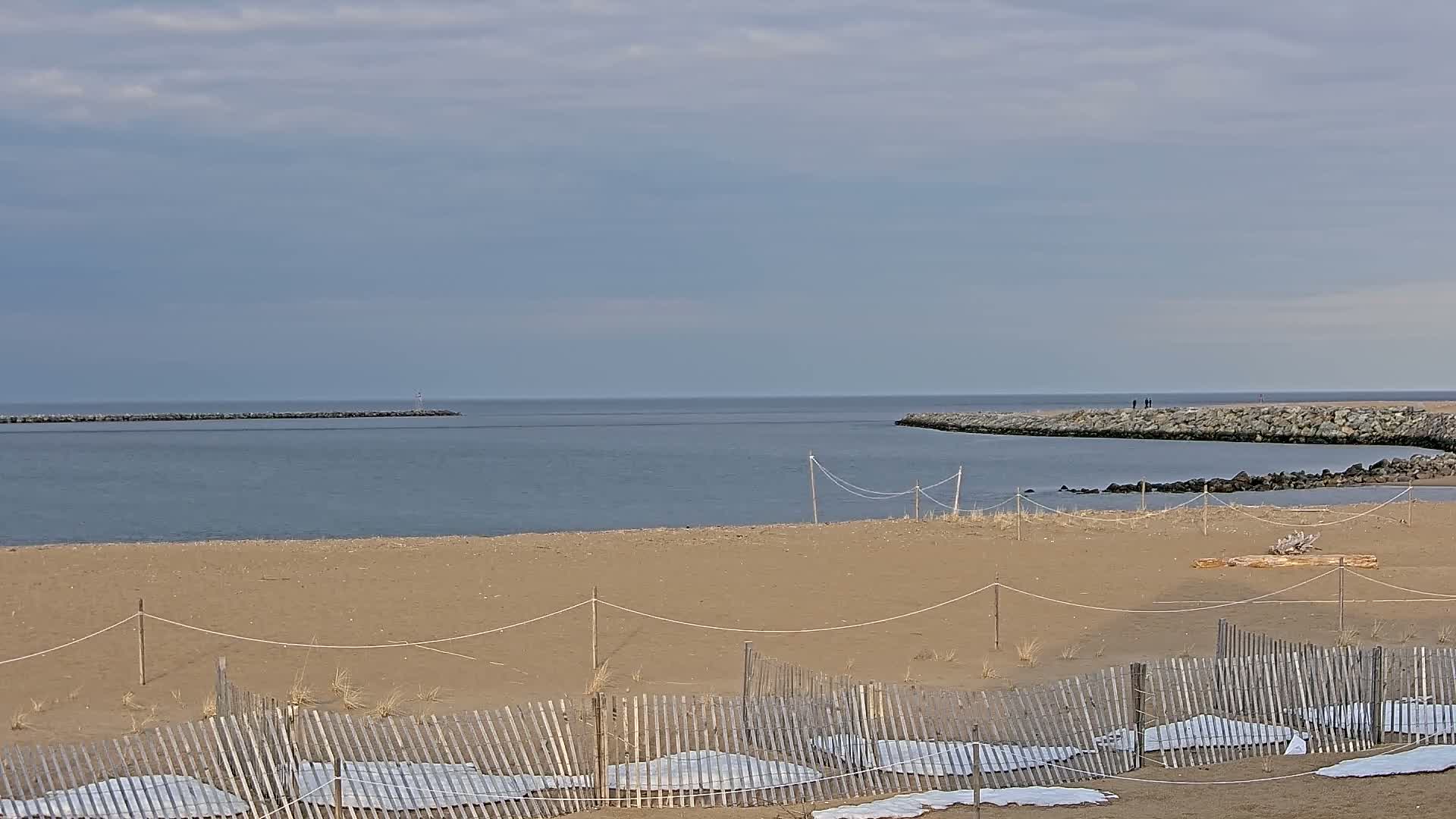A sandy beach with a wooden fence and coverings on the sand, overlooks a calm bay partially enclosed by rock jetties under a cloudy sky.