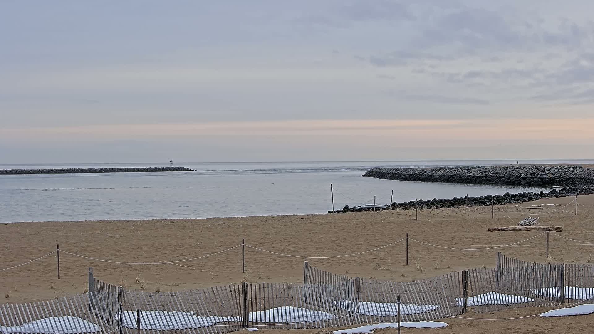 A sandy beach with a wooden fence and patches of snow is seen, leading to calm water and rock jetties under a mostly cloudy sky.