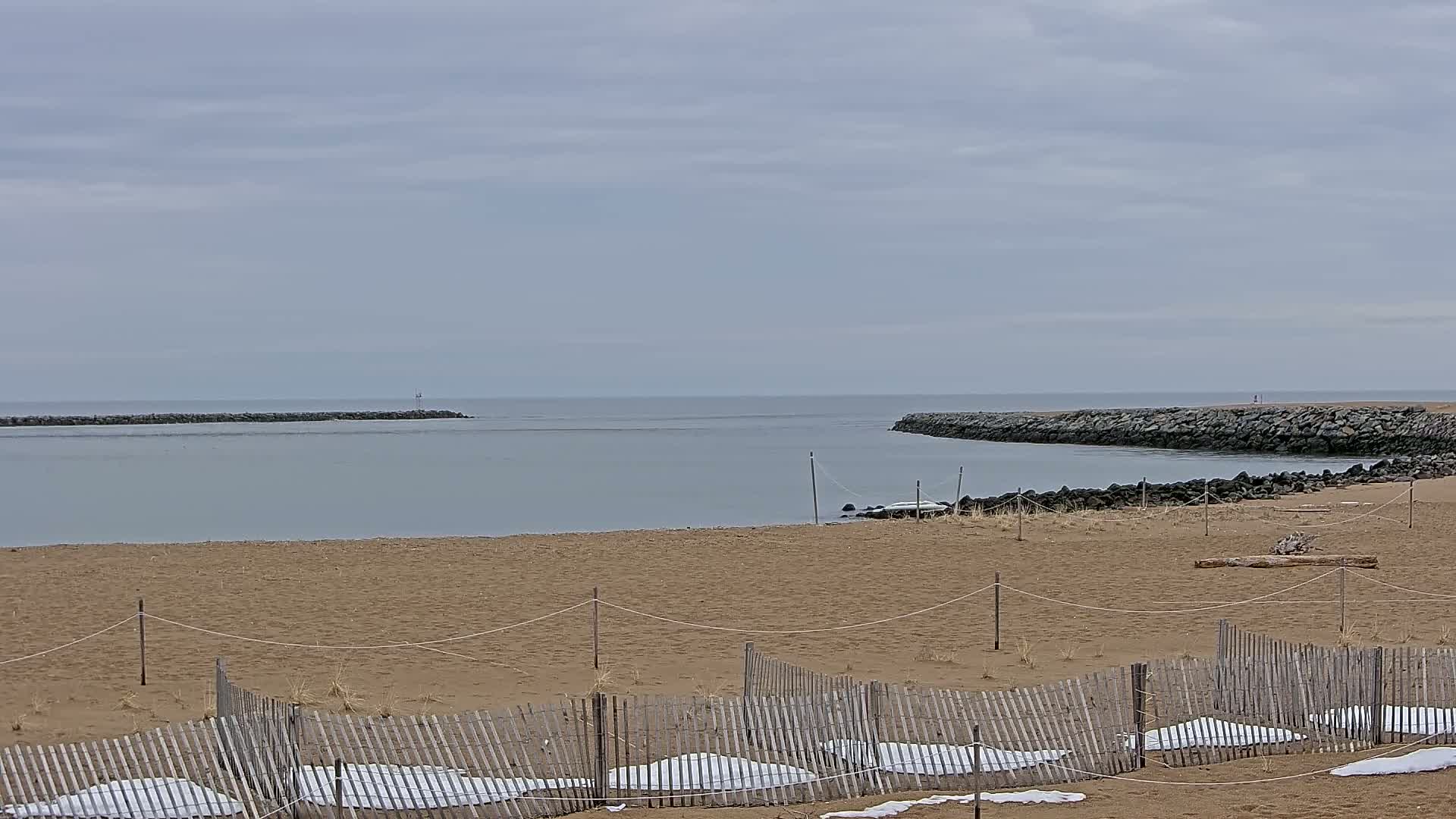 A sandy beach with a wooden fence partially covered in snow overlooks a calm body of water bordered by rock jetties under a cloudy sky.