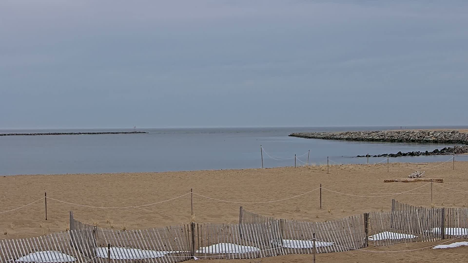 A sandy beach with a low wooden fence and patches of snow is seen under an overcast sky, with calm water and rock jetties in the background.