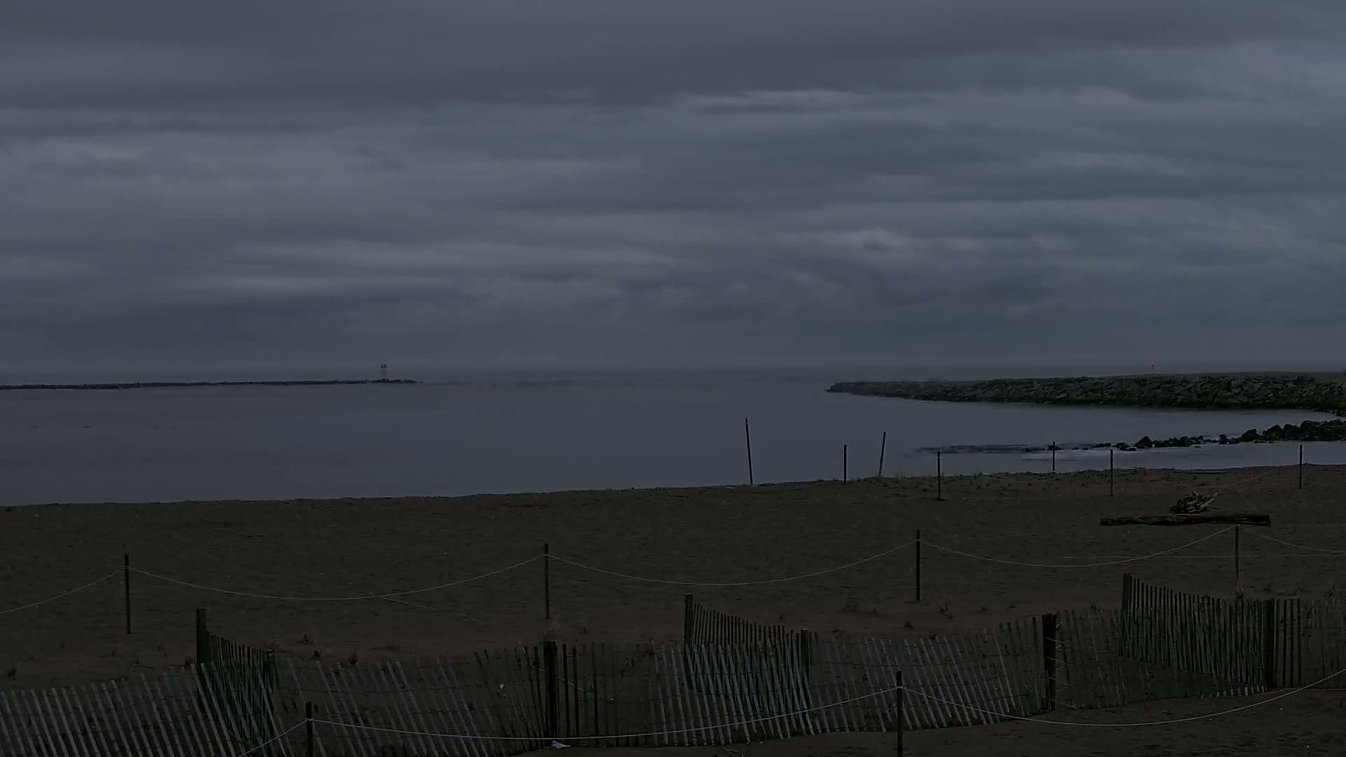 A dark, overcast sky hangs over a calm body of water, partially enclosed by rock jetties, where a sandy beach is bordered by a simple wooden fence.