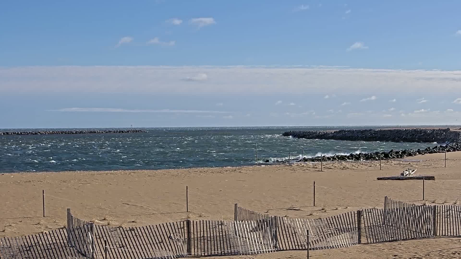 A sandy beach with a partially damaged wooden fence stretches toward a choppy sea with rock jetties under a partly cloudy sky.