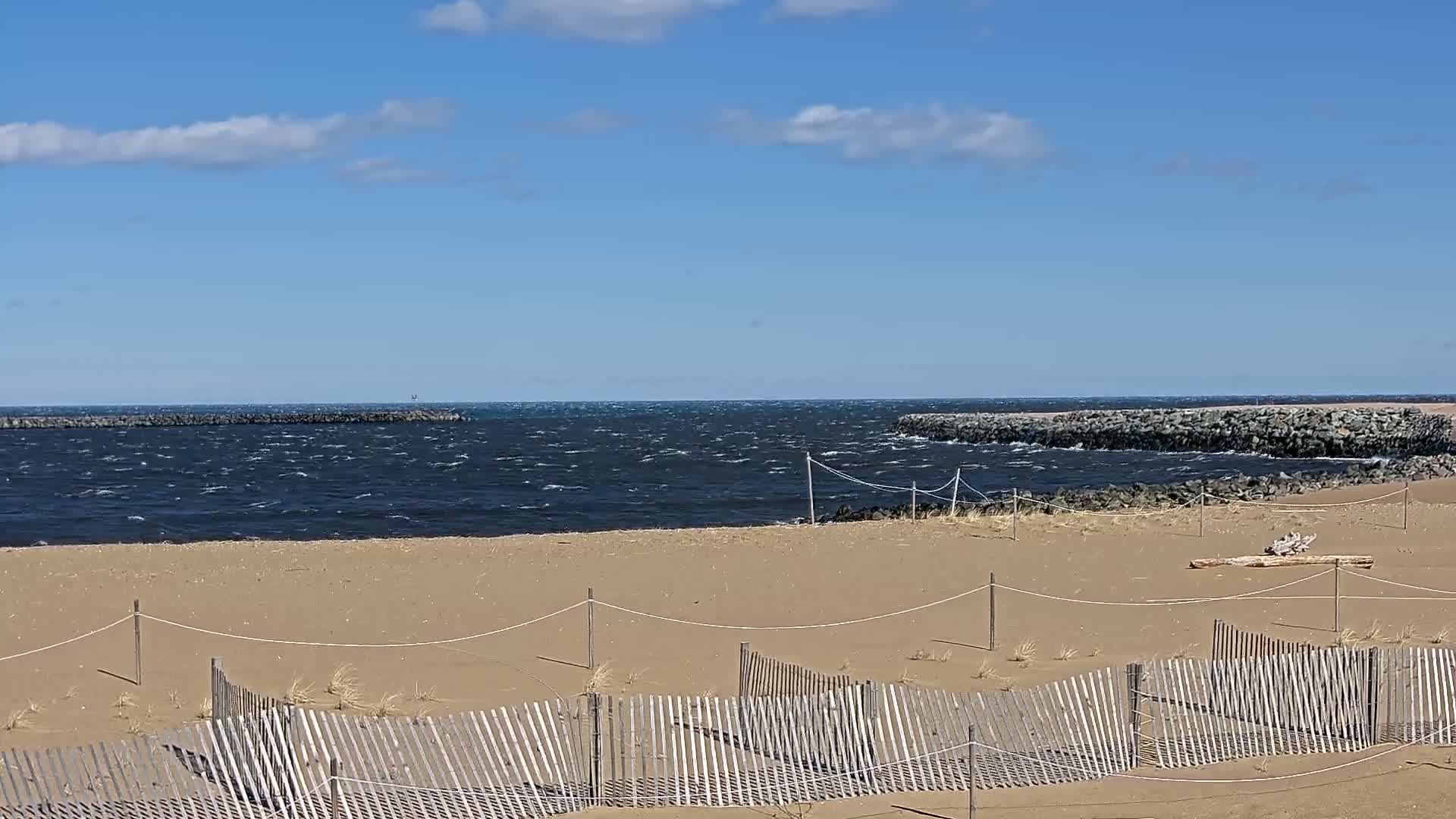 A sandy beach with a low wooden fence stretches toward a choppy, dark blue body of water partially enclosed by rock barriers under a partly cloudy blue sky.