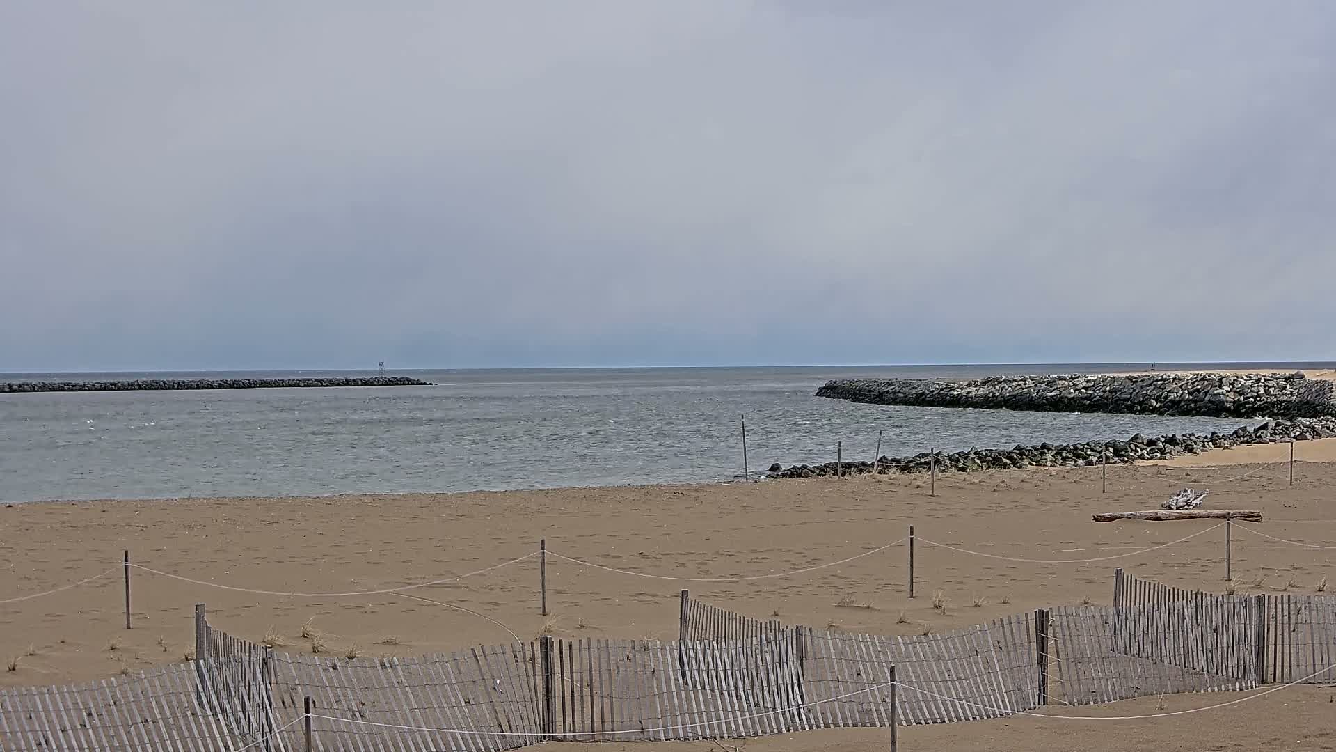 A sandy beach with a weathered wooden fence is situated between two rock jetties, under an overcast sky, overlooking a calm body of water.