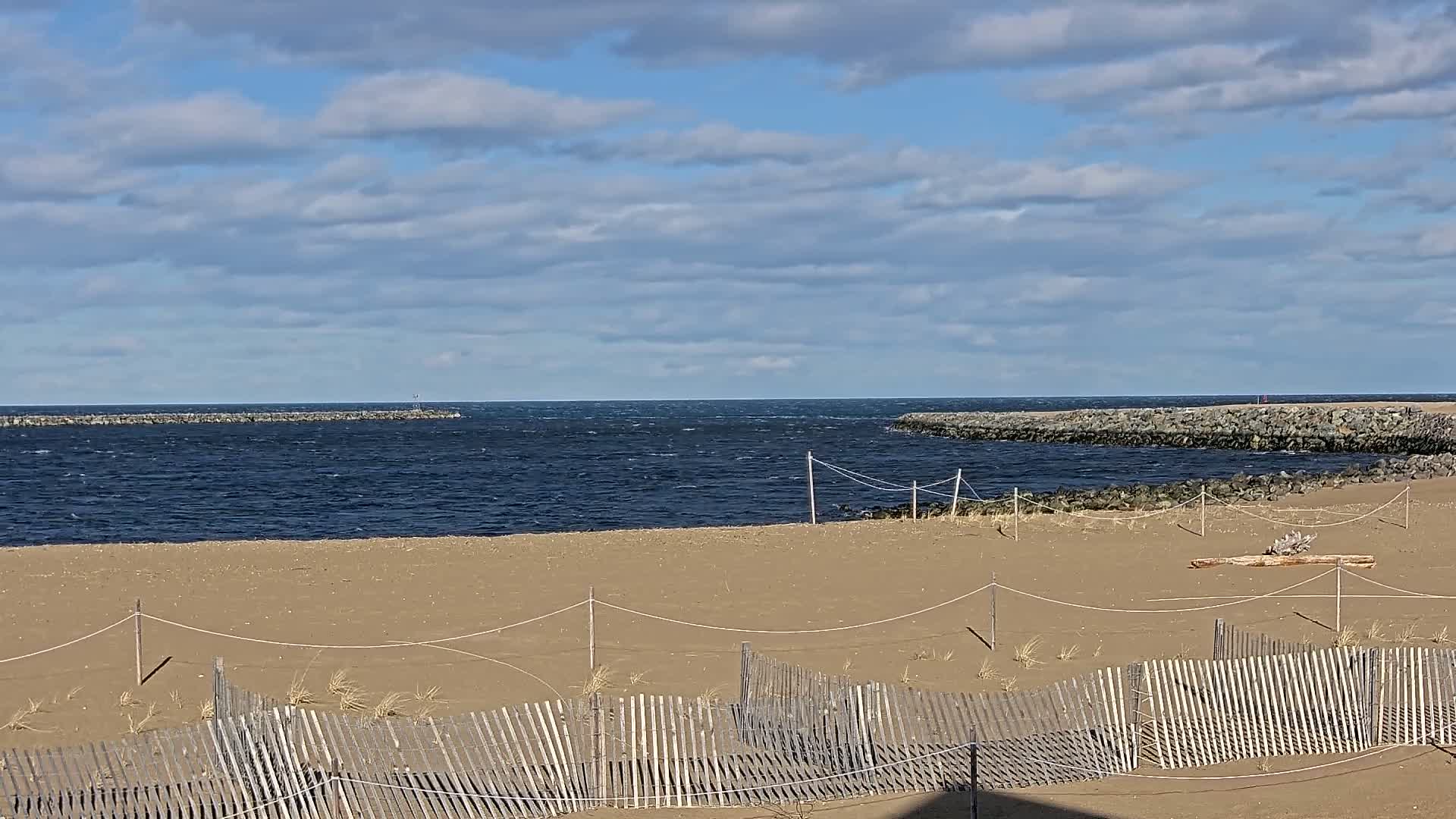 A sandy beach with a wooden fence is situated before a choppy ocean, partially enclosed by rock jetties under a partly cloudy sky.