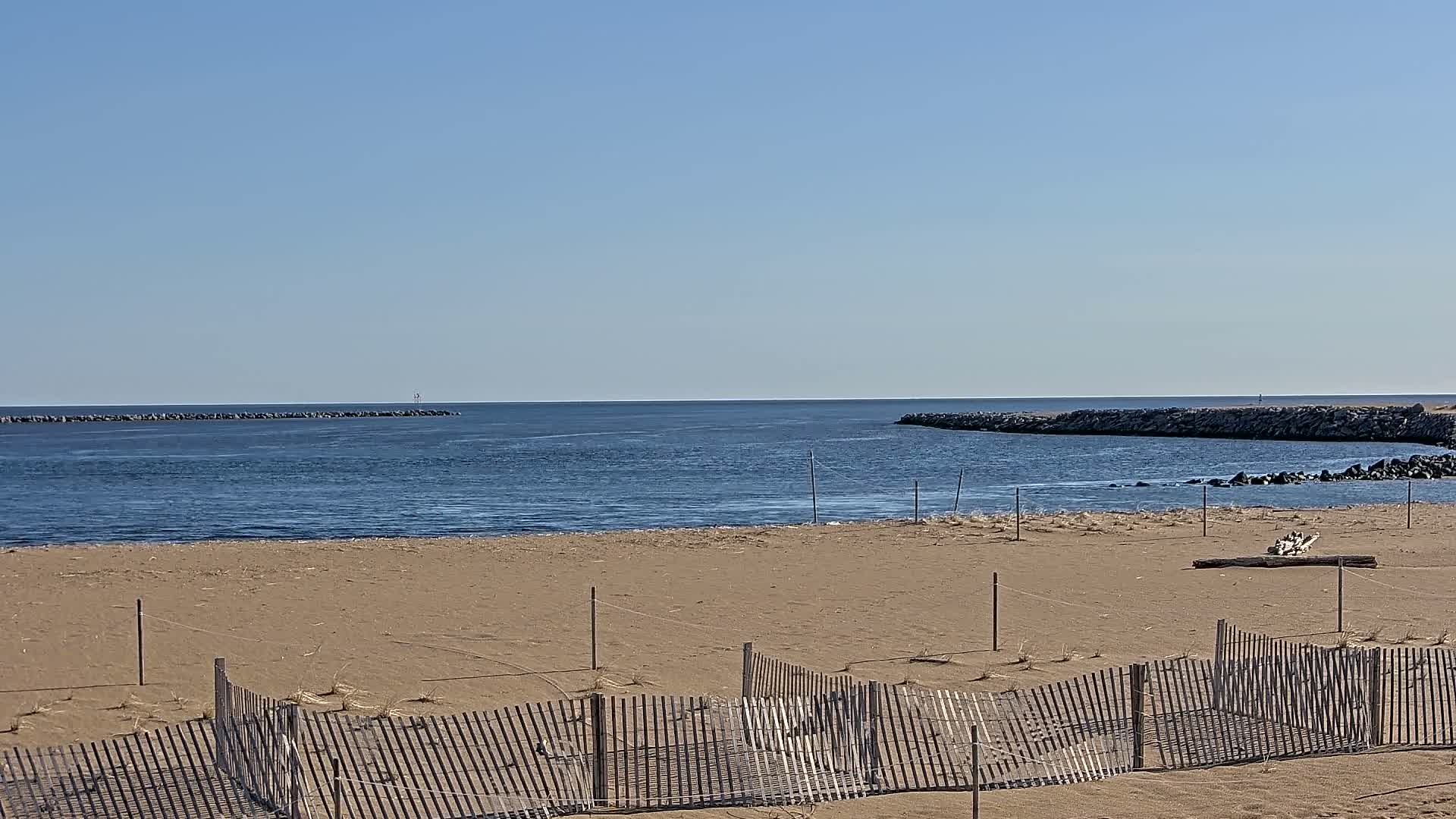 A sandy beach with a wooden fence stretches toward a calm, blue body of water bordered by rock jetties under a clear, sunny sky.
