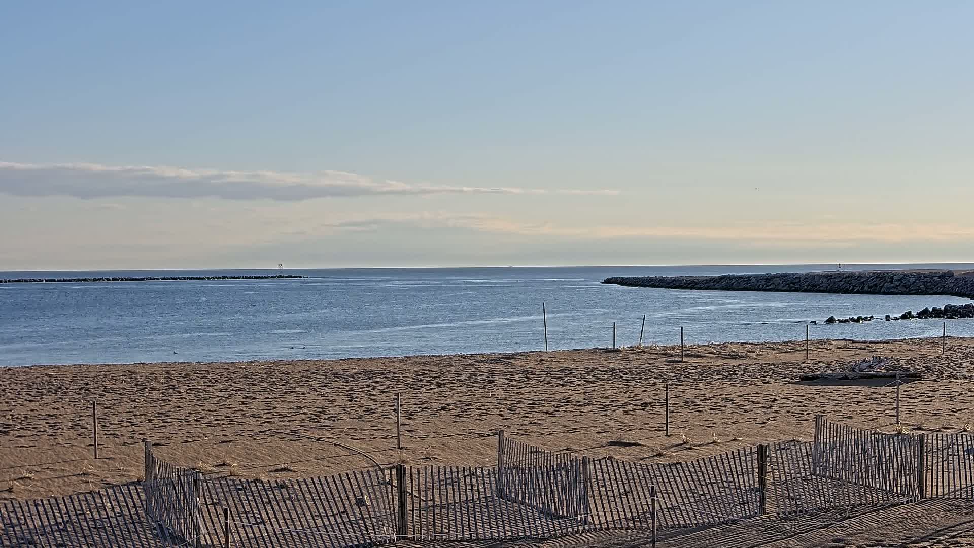 A sandy beach with a calm, blue ocean under a mostly clear sky, featuring a series of low fences in the foreground.