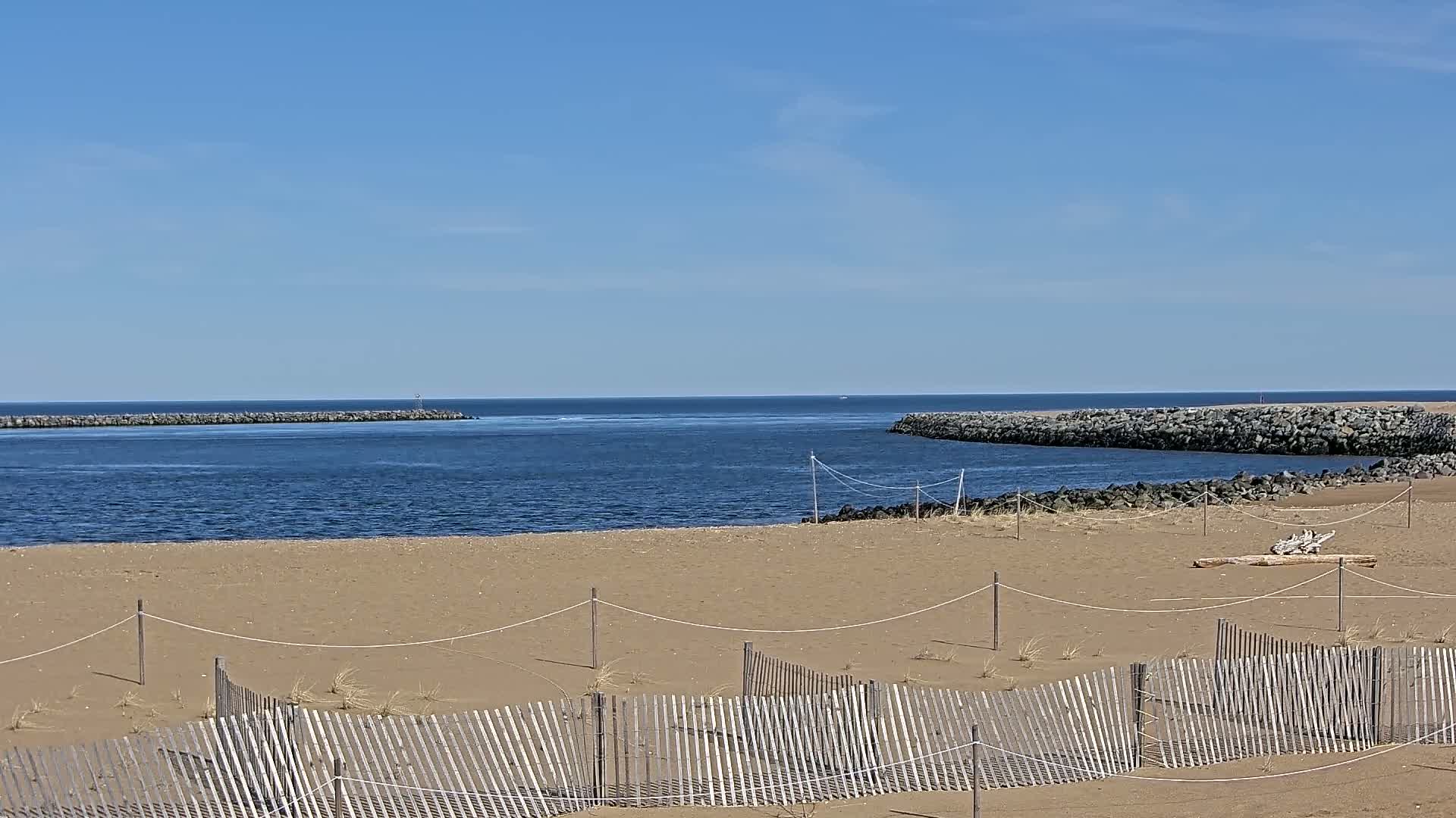 A sandy beach with a wooden fence is visible in the foreground, overlooking a calm blue body of water, bordered by rock jetties under a clear blue sky.