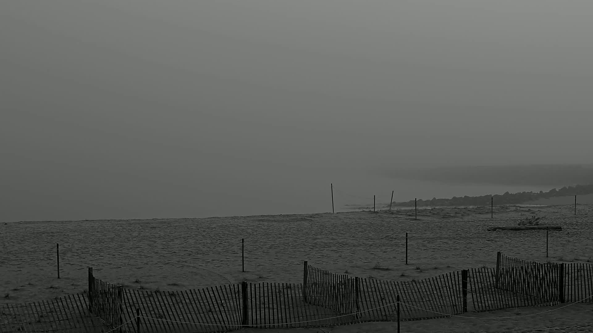 A foggy beach scene shows a sandy shore with short wooden fences, leading to a hazy ocean.