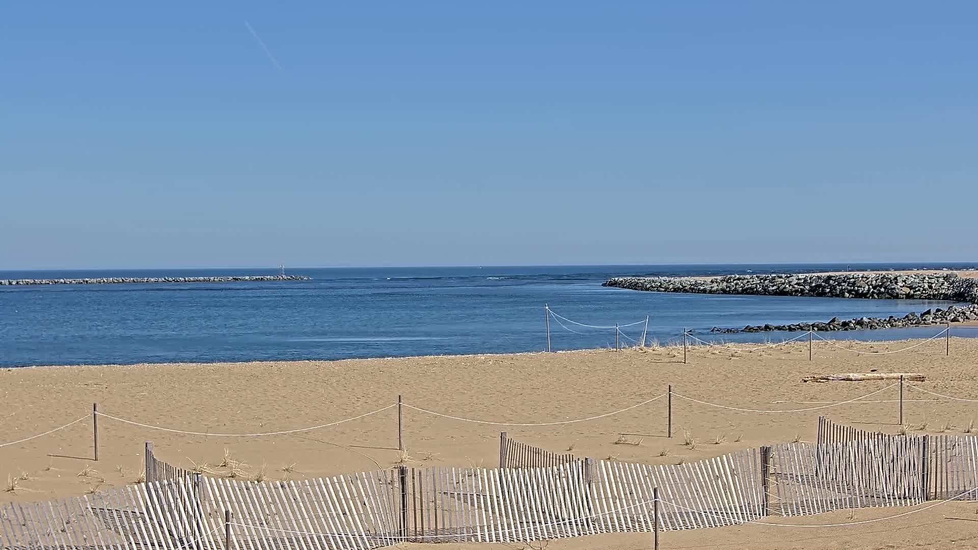 A sandy beach with a low wooden fence stretches towards a calm body of water under a clear blue sky.