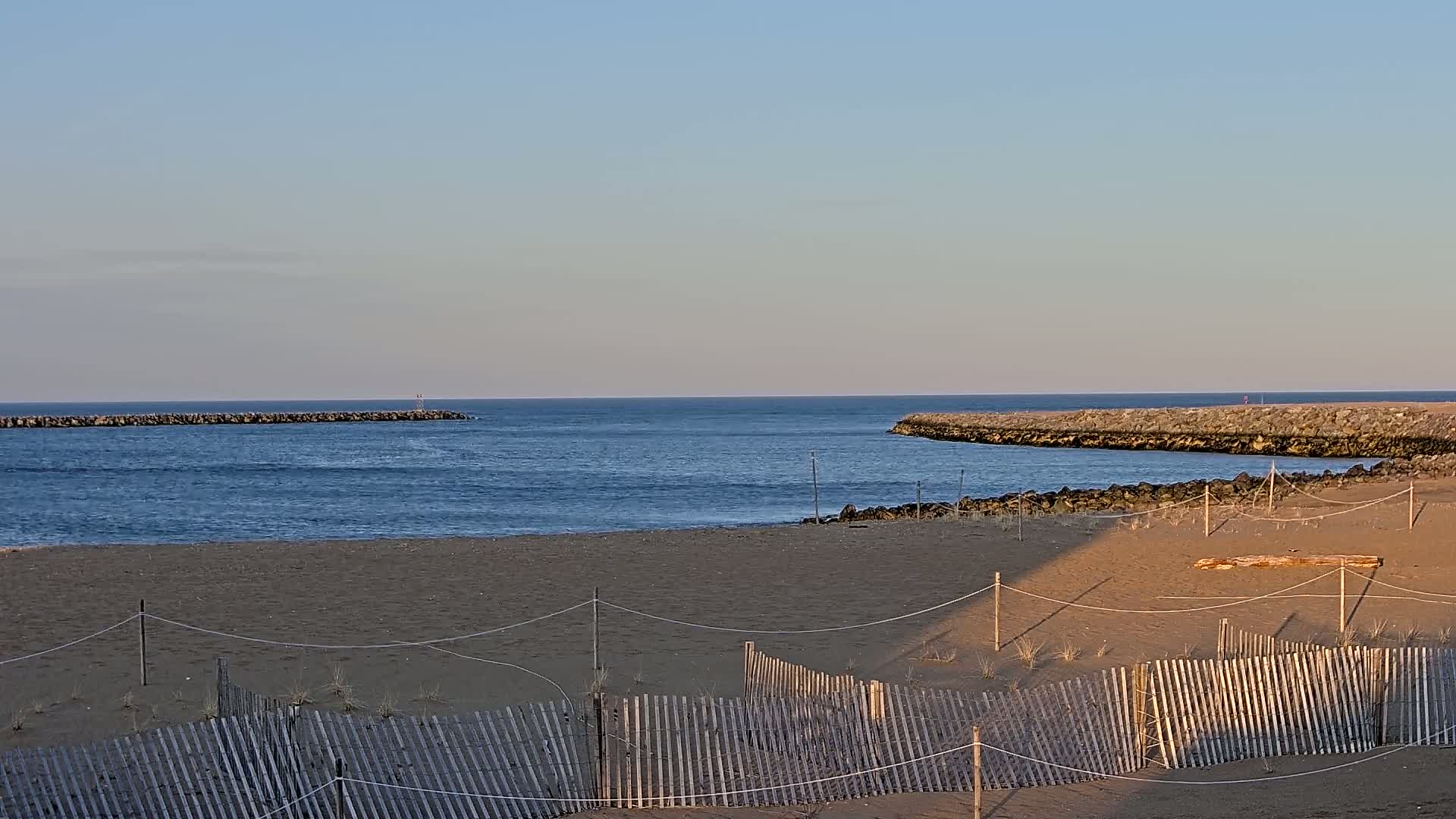 A sandy beach with a wooden fence stretches towards a calm ocean inlet bordered by rock jetties under a clear, pale blue sky.