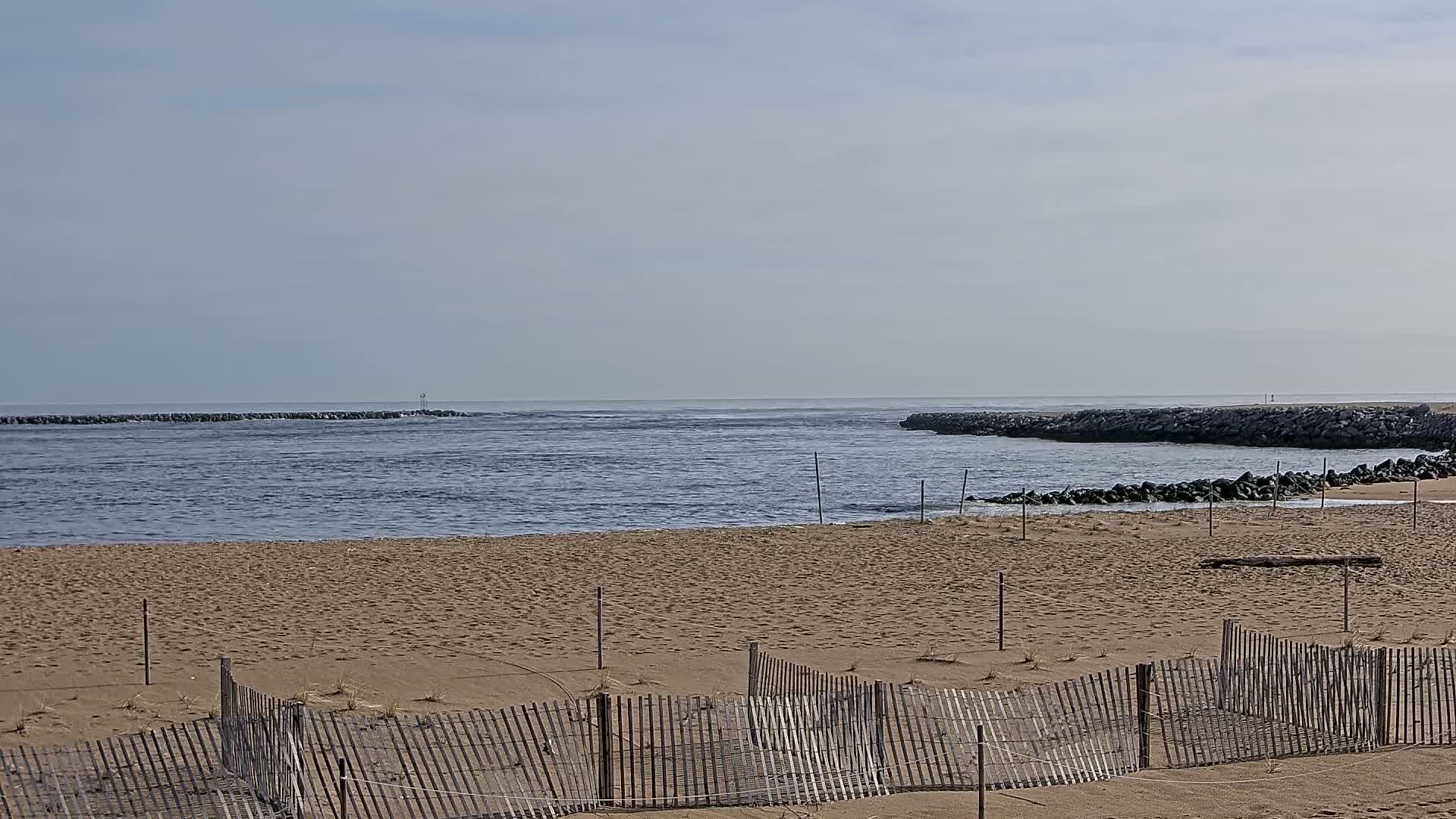 A sandy beach with a weathered wooden fence stretches toward a calm ocean inlet bordered by rock jetties under a clear, bright sky.