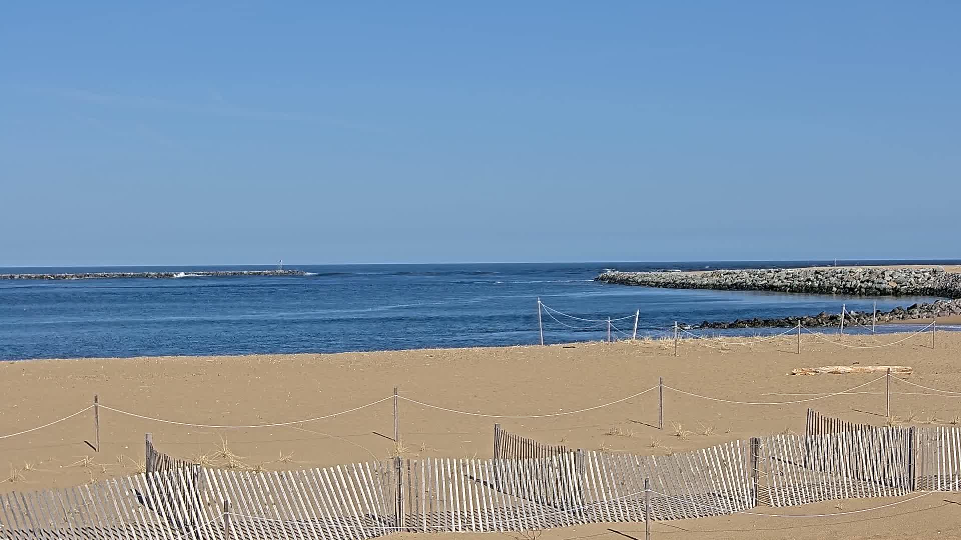 A sandy beach with a wooden fence stretches towards a calm blue ocean, partially enclosed by rock jetties under a clear blue sky.