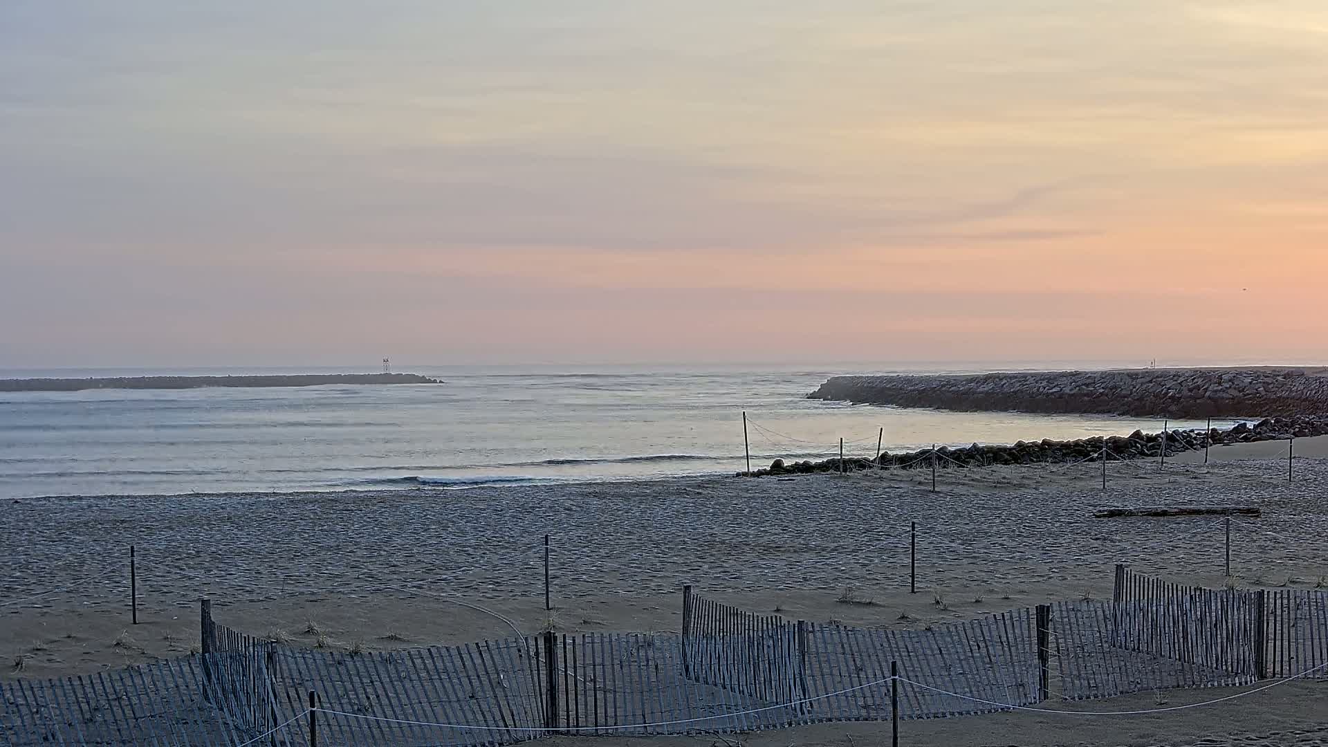 A sandy beach with a low wooden fence stretches toward a calm ocean inlet at sunrise under a pale pink sky.