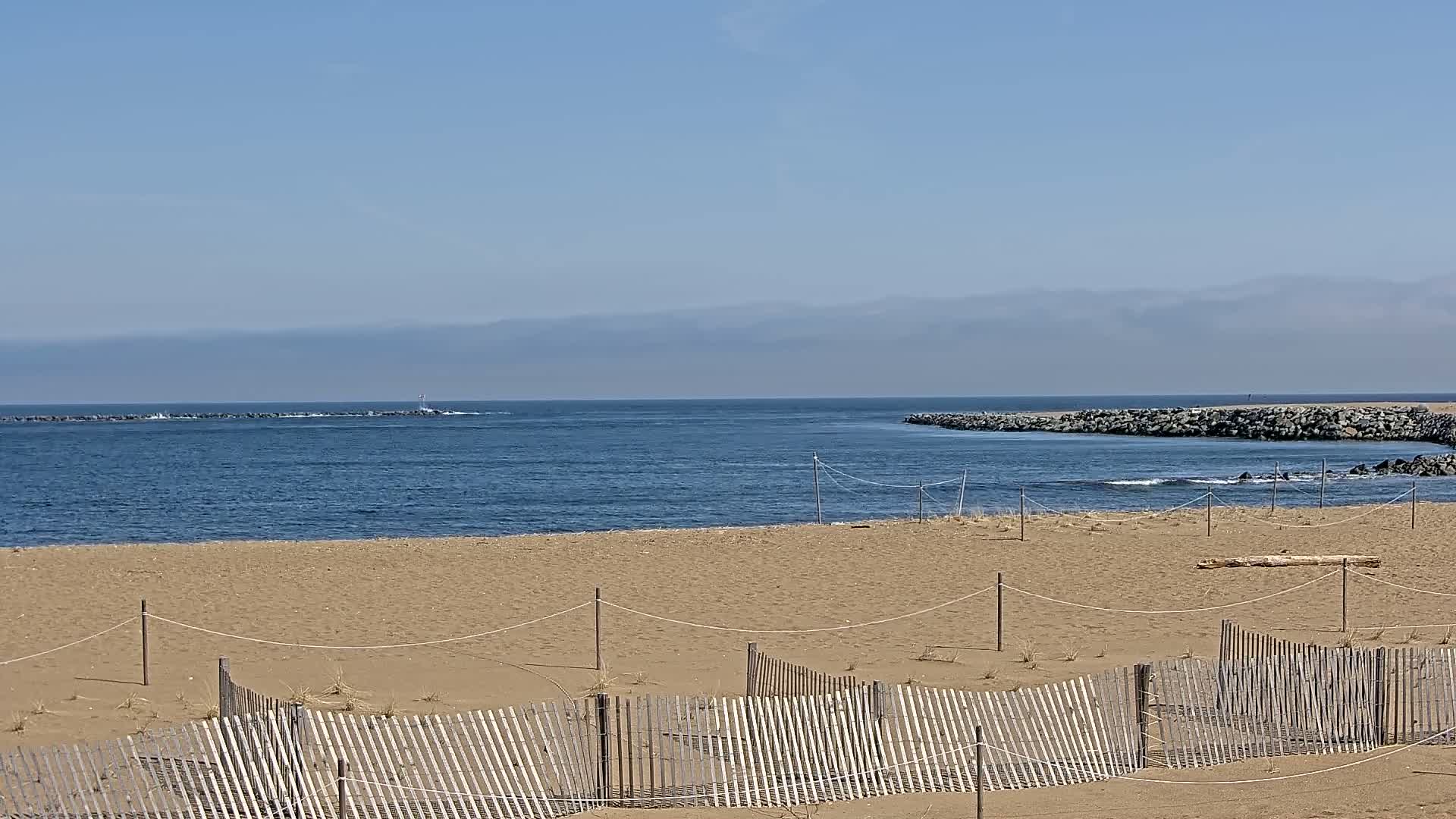 A sandy beach with a wooden fence in the foreground overlooks a calm ocean inlet on a clear day.
