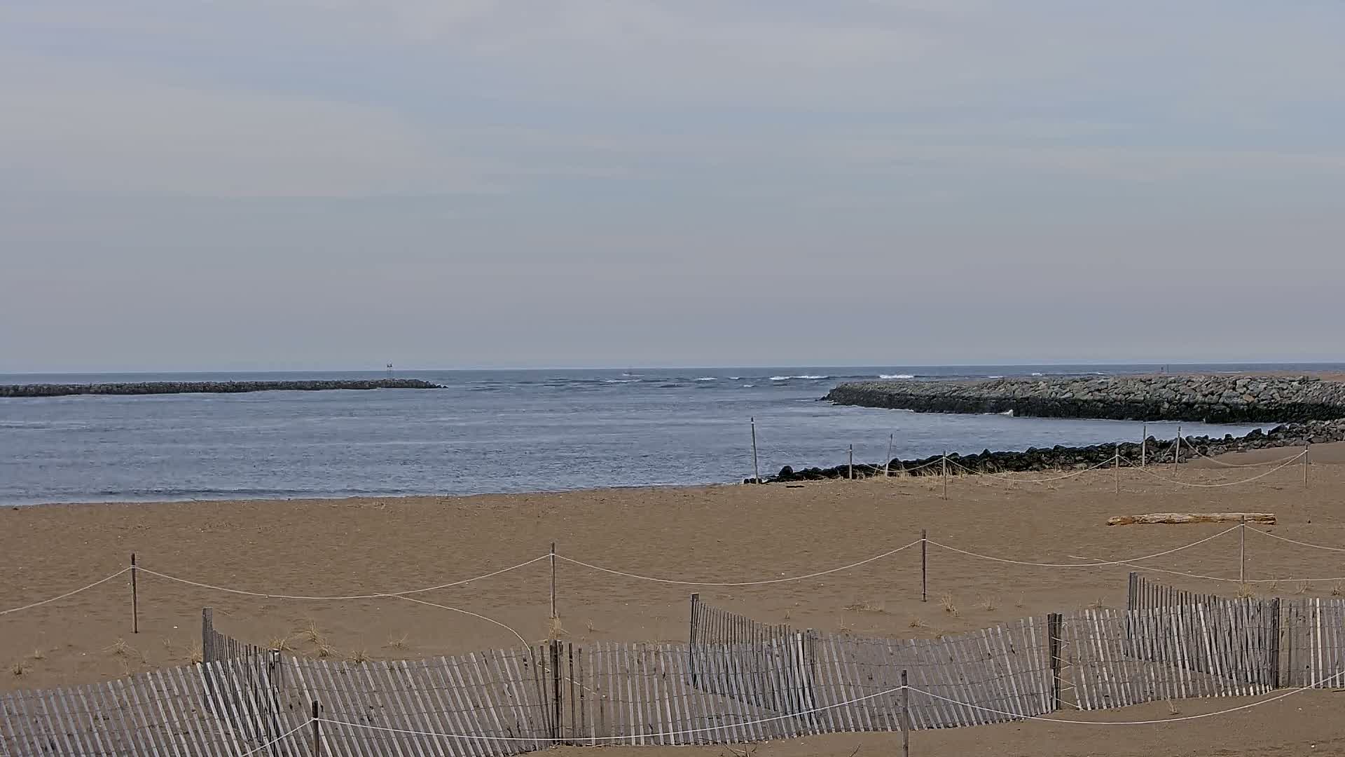 A sandy beach with a wooden fence stretches towards a calm ocean inlet bordered by rock jetties under a cloudy sky.
