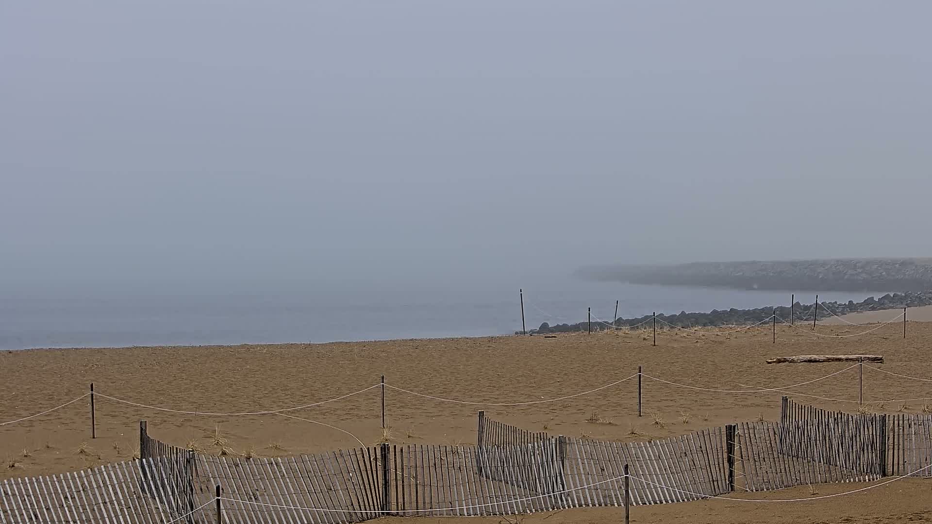 A sandy beach with a low wooden fence stretches towards a foggy, calm ocean meeting a dark rock jetty.
