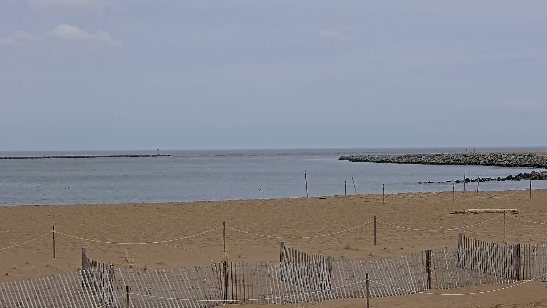 A sandy beach with a wooden fence stretches towards a calm, gray body of water bordered by rock jetties under an overcast sky.