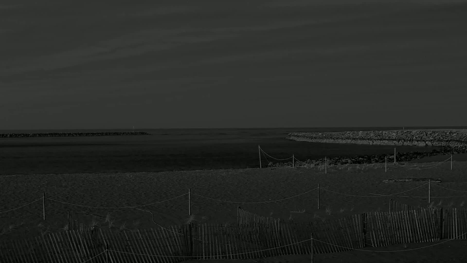 A dark, sandy beach with a low wooden fence is situated between two rocky jetties under a dusky sky.