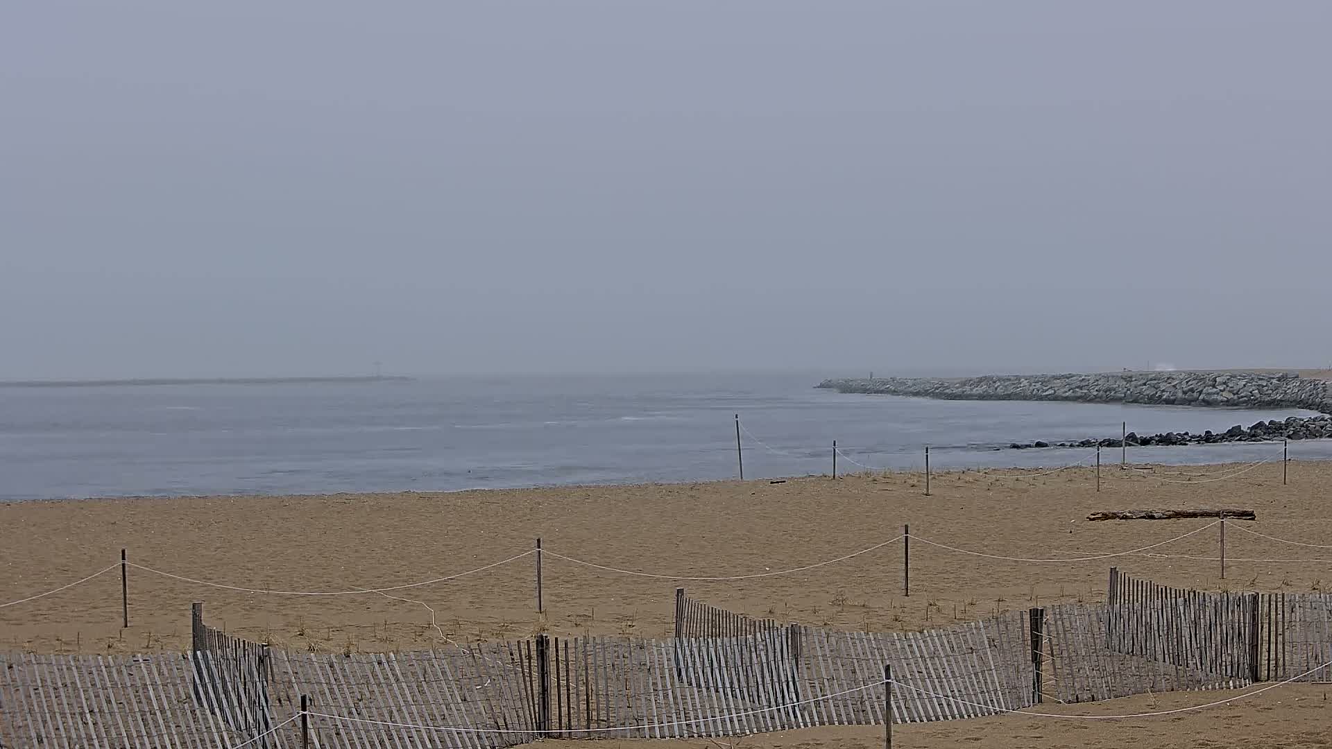 A sandy beach with low wooden fences is visible, leading to a calm, foggy ocean meeting a rocky jetty.